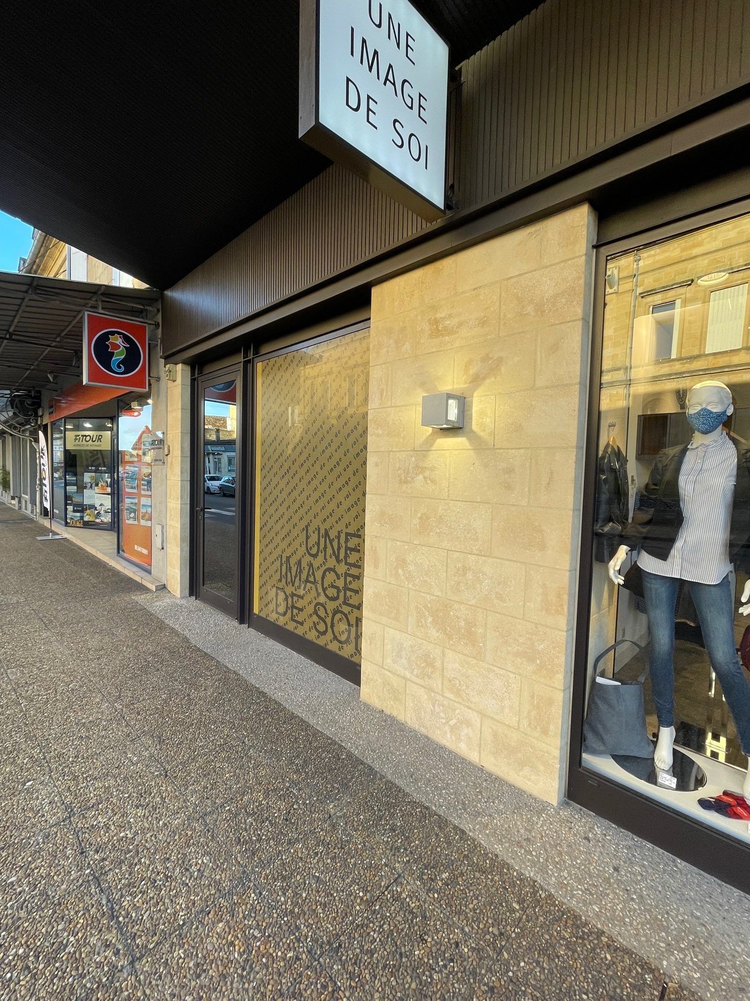 Extérieur d'une boutique de vêtements. Façade beige clair avec vitrine présentant un mannequin en jean et haut rayé.