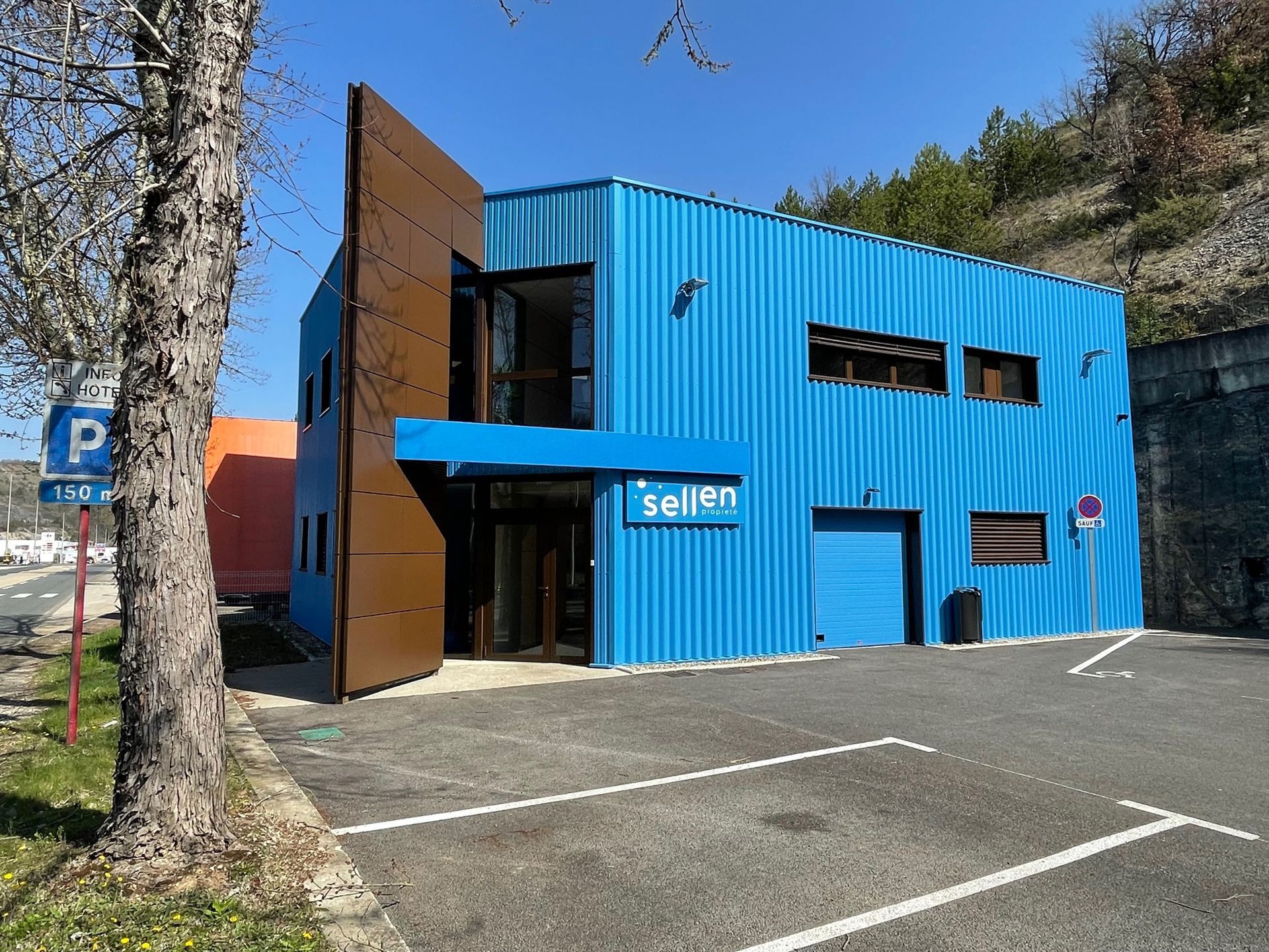 Bâtiment moderne bleu et marron avec parking, arbre et montagne en arrière-plan.