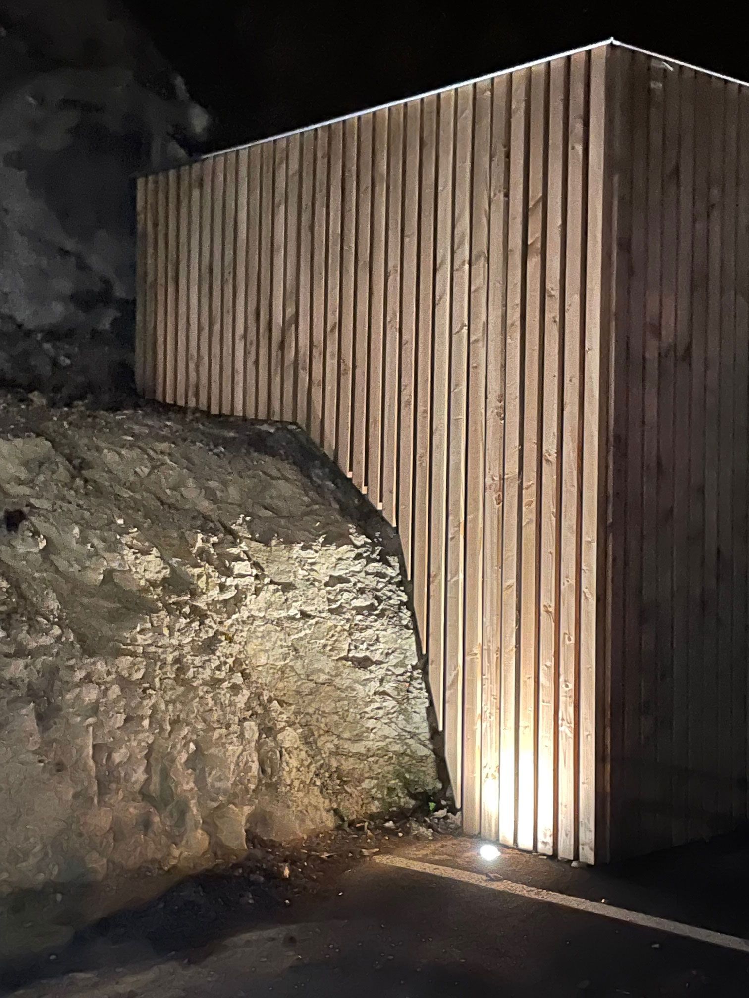 Un mur en bois éclairé jouxte une formation rocheuse. La scène est sombre, avec une douce lumière sur le bois.