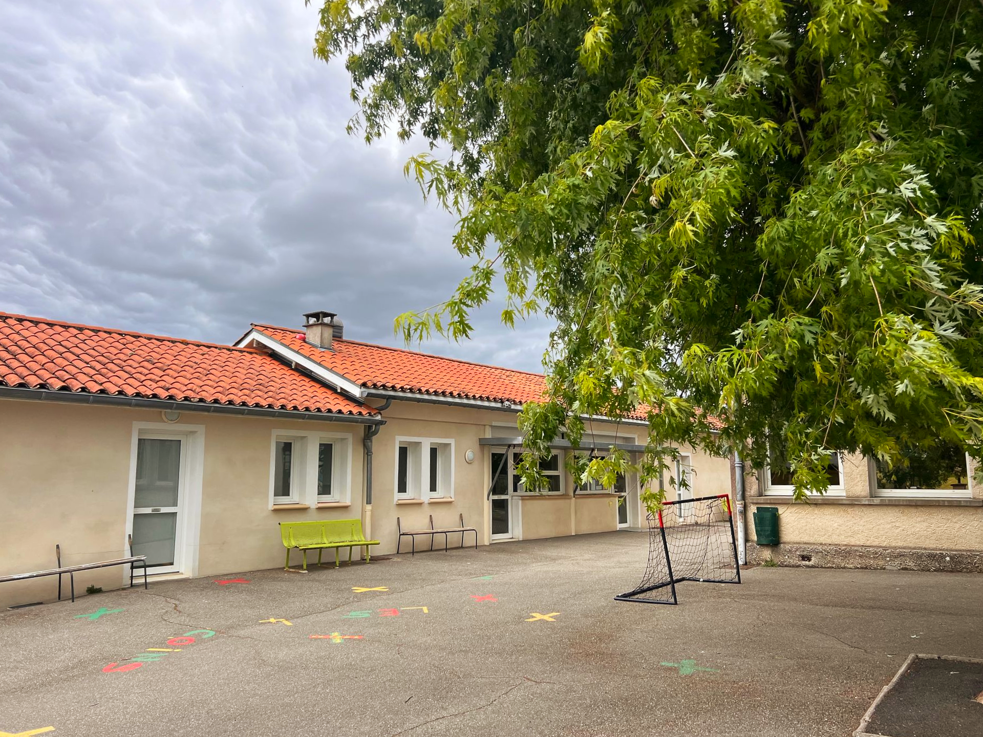 Bâtiment au toit de tuiles, aux murs blancs et aux poutres de soutien colorées. Un banc vert est installé devant.