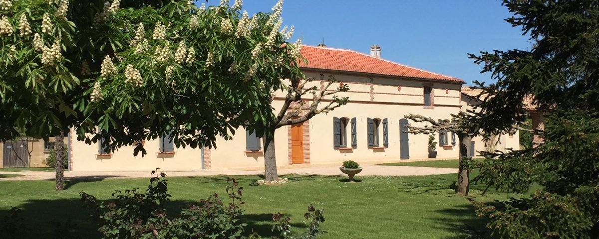 Un bâtiment clair au toit de tuiles rouges, derrière des arbres et de l'herbe verte, par une journée ensoleillée.