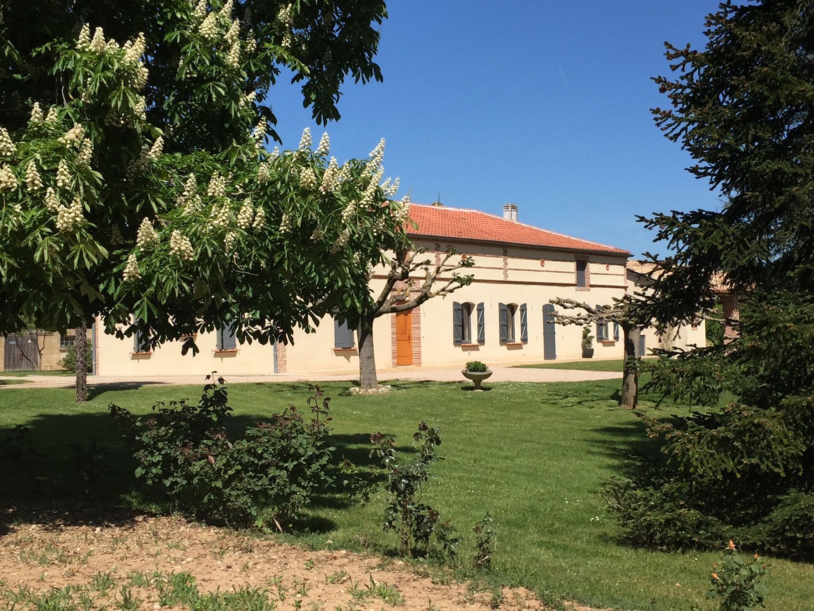 Bâtiment beige au toit de tuiles rouges, encadré d'arbres et de pelouse verte, sous un ciel bleu.