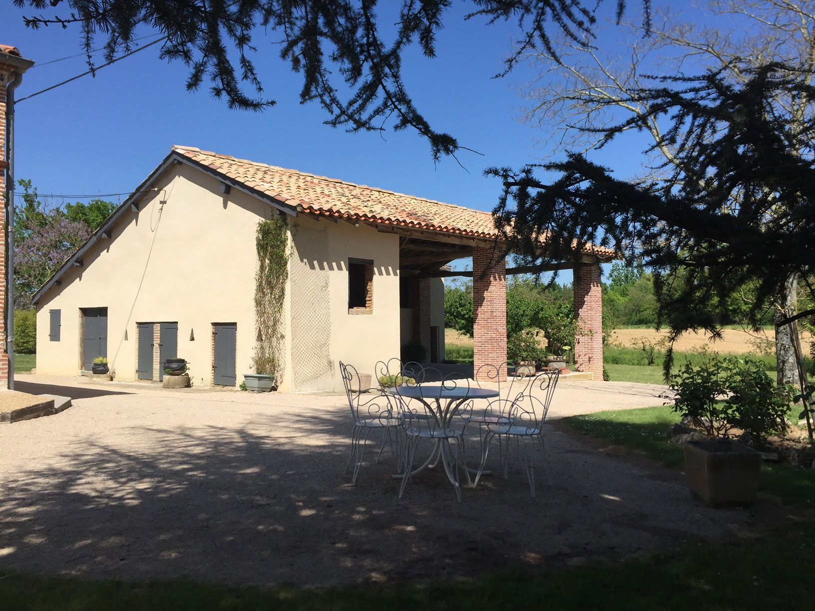 Bâtiment en pierre avec toit en tuiles de terre cuite, terrasse extérieure et cadre ensoleillé et arboré.