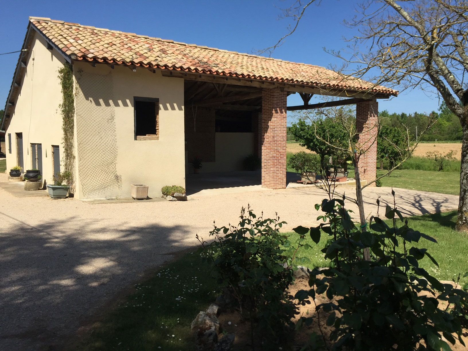 Bâtiment de couleur beige avec un toit de tuiles en terre cuite, un porche ouvert et une allée de gravier dans un cadre rural ensoleillé.