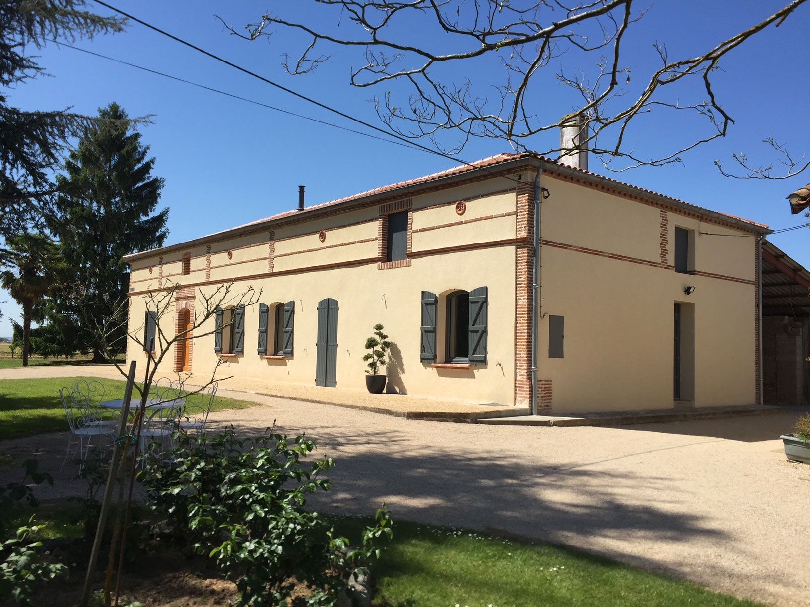 Ferme beige avec toit de tuiles rouges, volets bleus et allée de gravier par une journée ensoleillée.