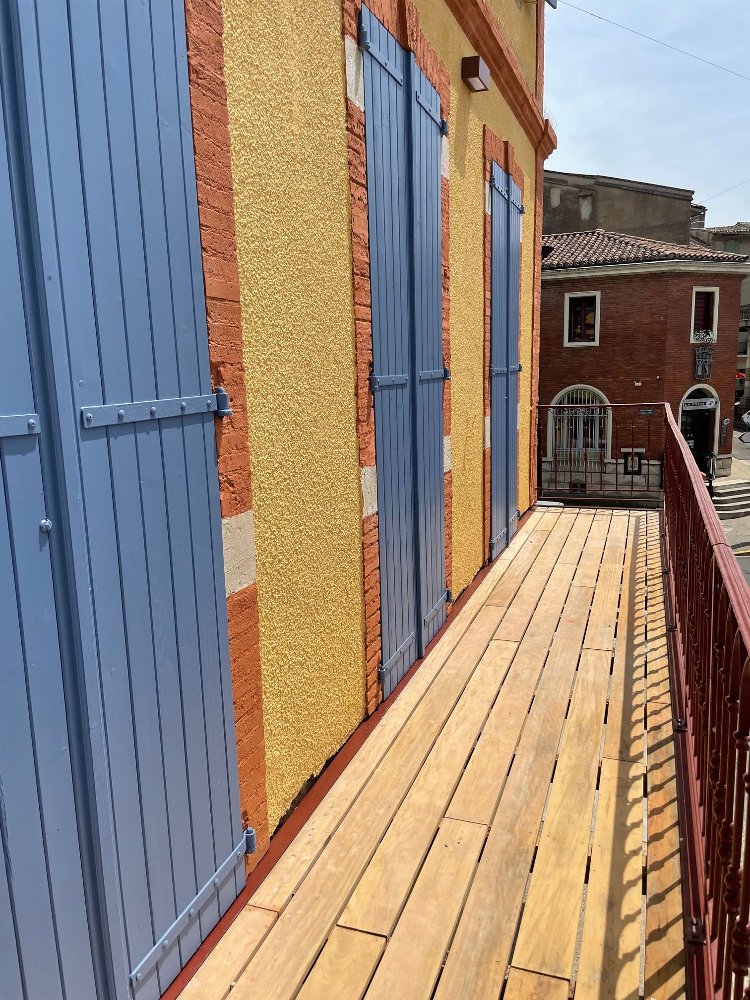 Balcon avec volets bleus, mur en stuc jaune, boiseries orange, rambarde rouge et parquet. Un immeuble en briques se dresse à l'arrière-plan.