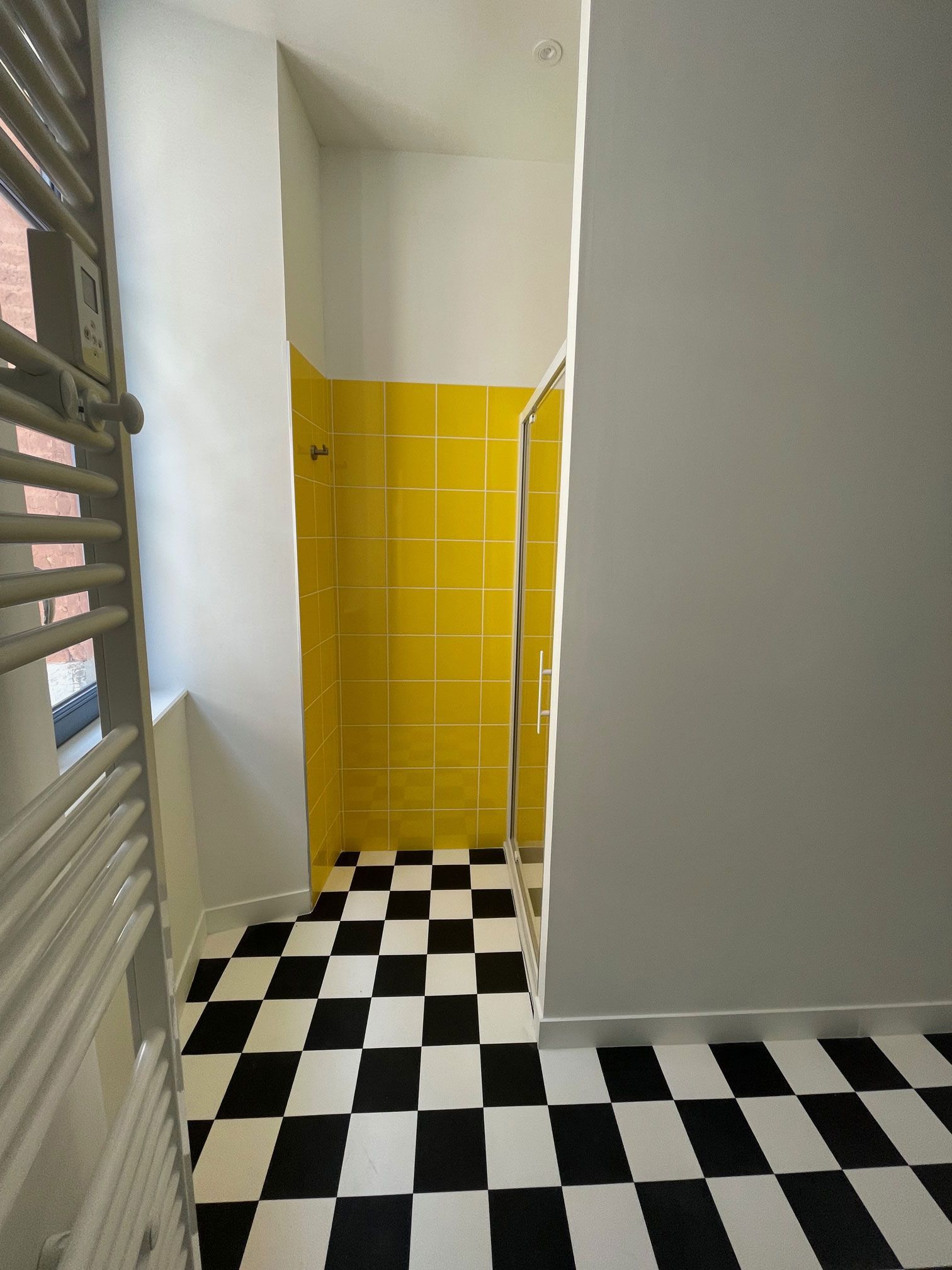 Douche carrelée de jaune dans une salle de bains avec sol à damier noir et blanc et mur gris.