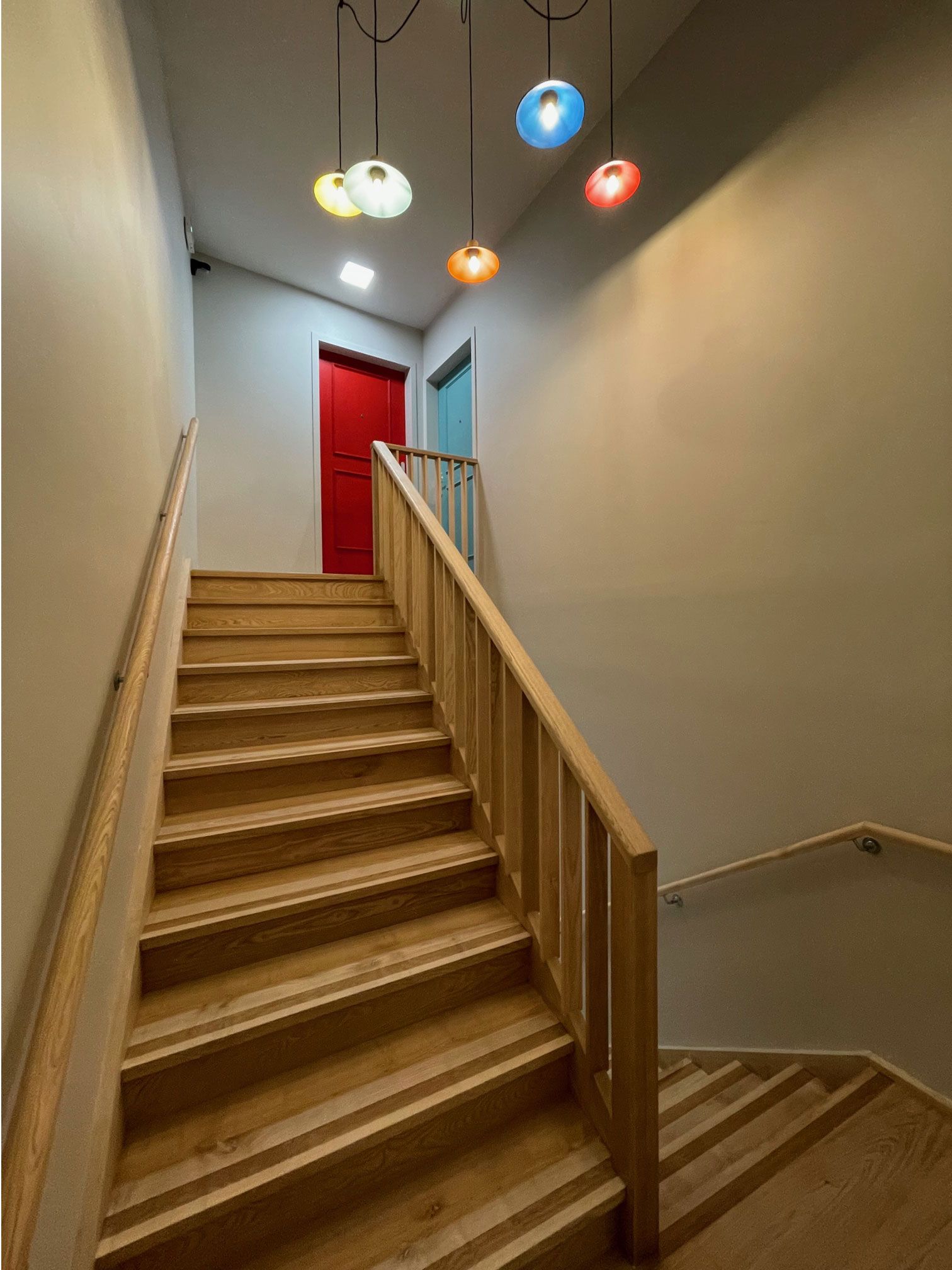 Un escalier en bois monte, orné de guirlandes lumineuses colorées et d'une porte rouge en haut.