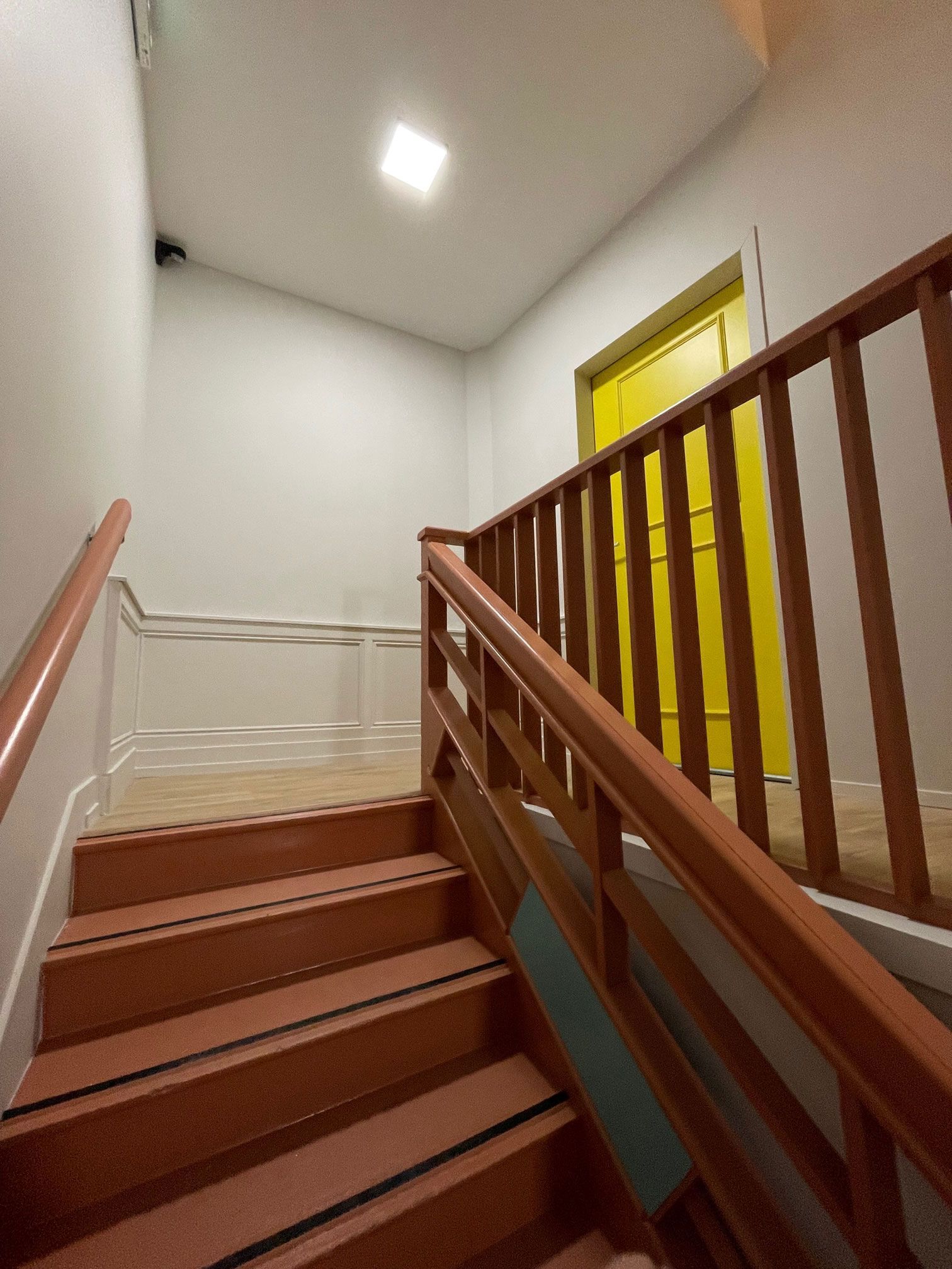 Escalier en bois menant à une porte jaune. Rambarde marron à droite, murs clairs, éclairage au plafond.