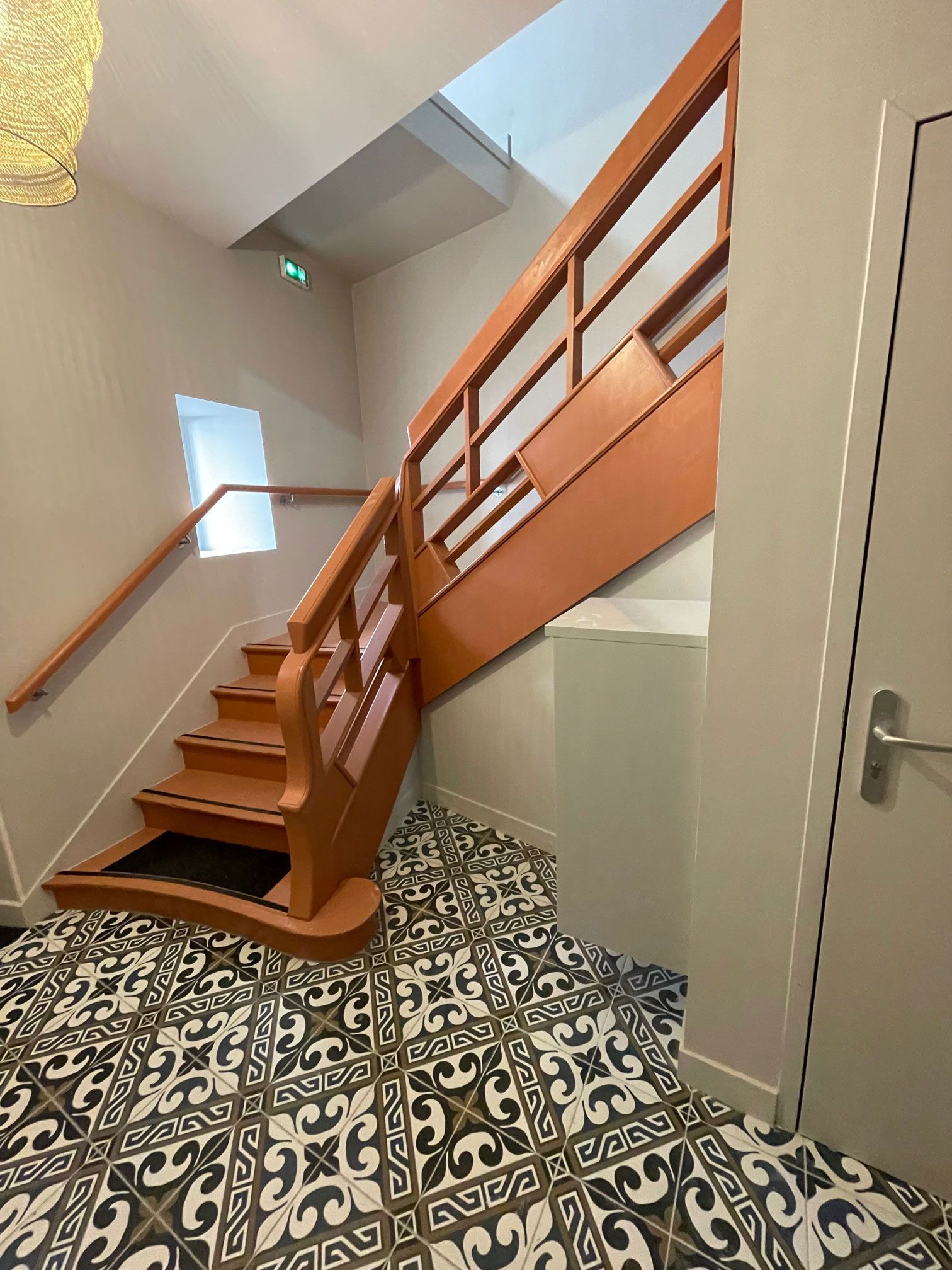 Escalier avec rampe et marches orange, sol carrelé à motifs et une porte sur la droite.