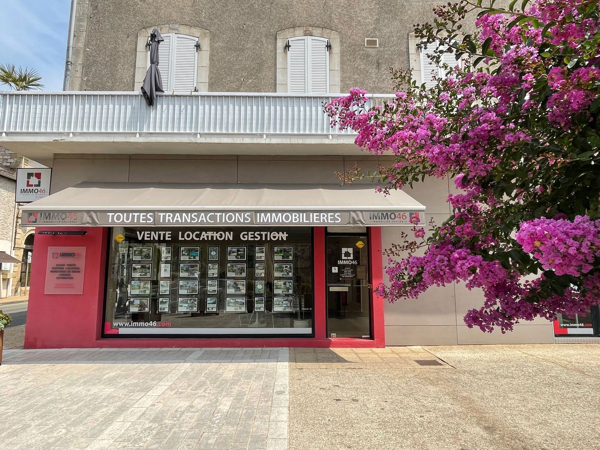 Agence immobilière avec vitrines, façade rose et fleurs violettes en pleine floraison.