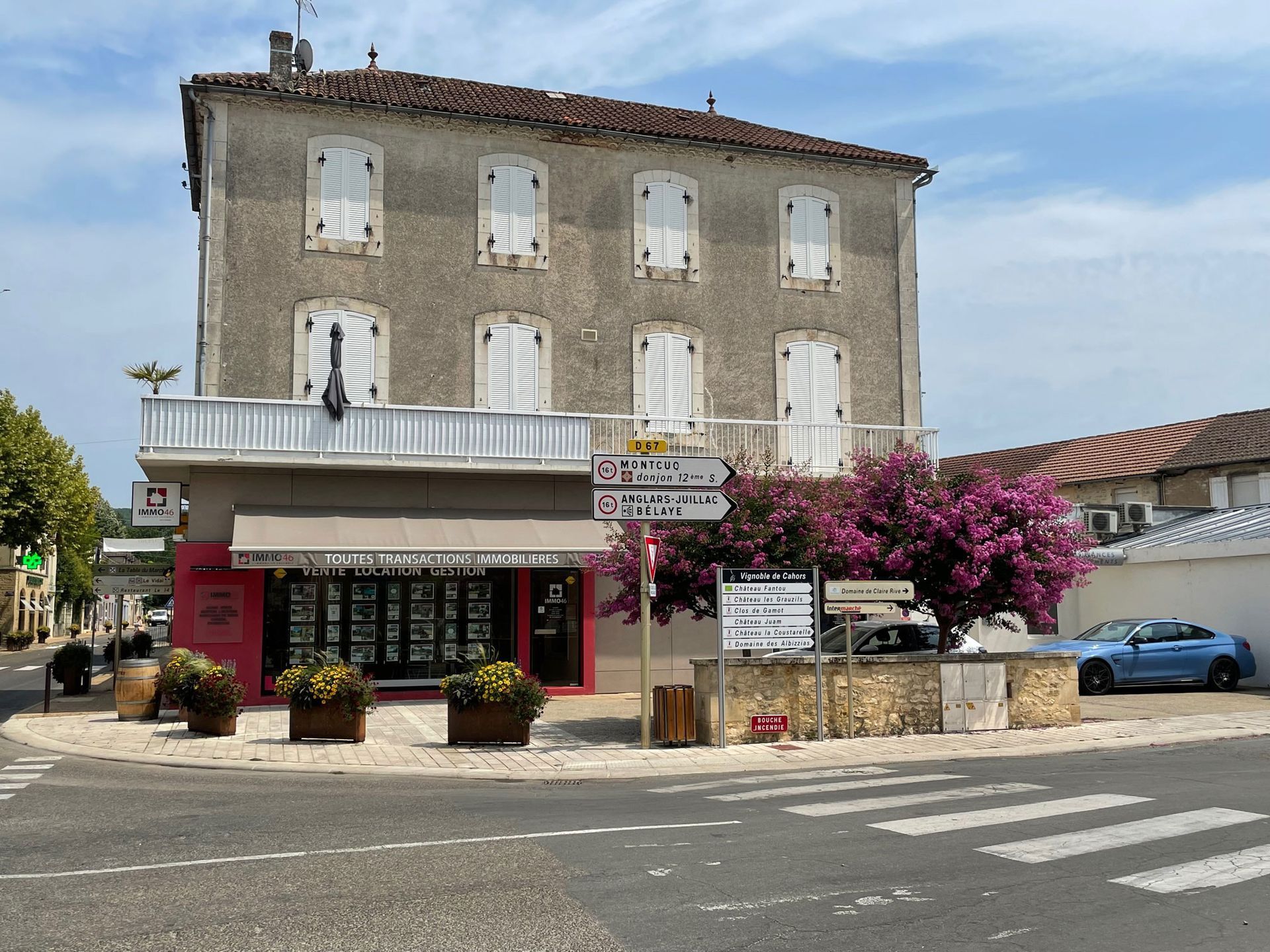 Bâtiment avec façade commerciale à un rond-point, avec une voiture bleue, des fleurs roses et un ciel bleu.