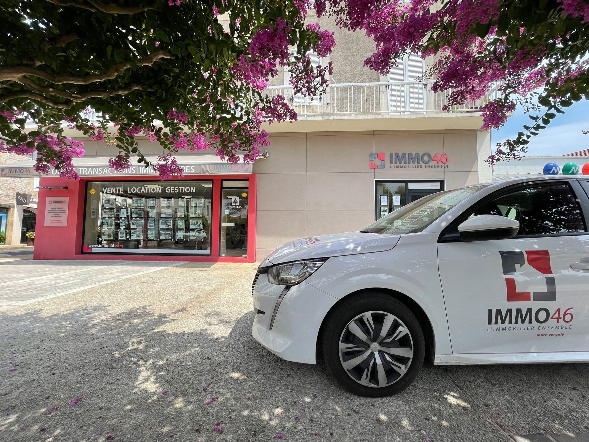 Voiture blanche avec le logo Immo46 garée devant une agence immobilière, sous un arbre en fleurs.