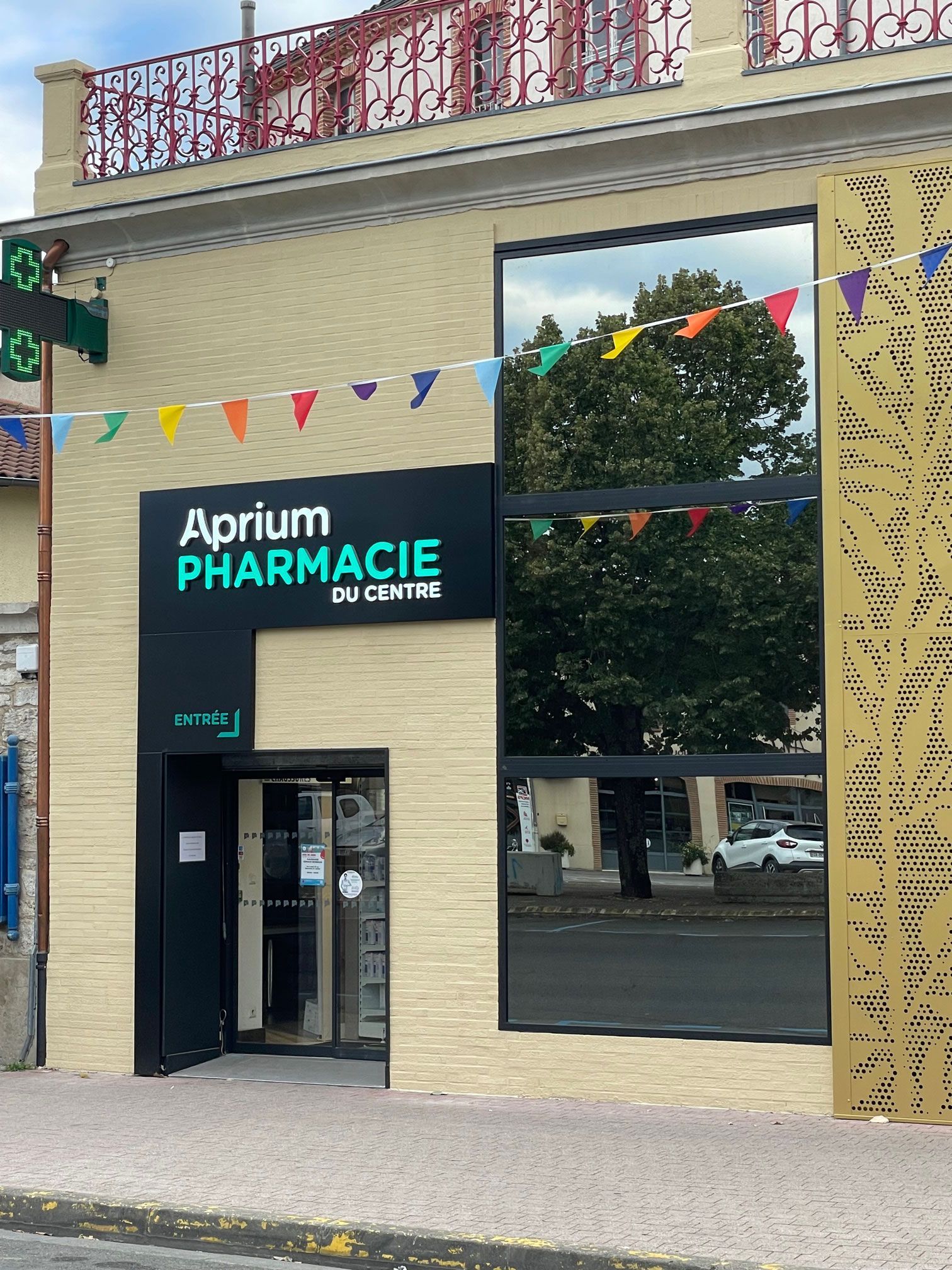 Façade de pharmacie avec enseigne turquoise et noire, grande vitrine reflétant des arbres et drapeaux colorés.
