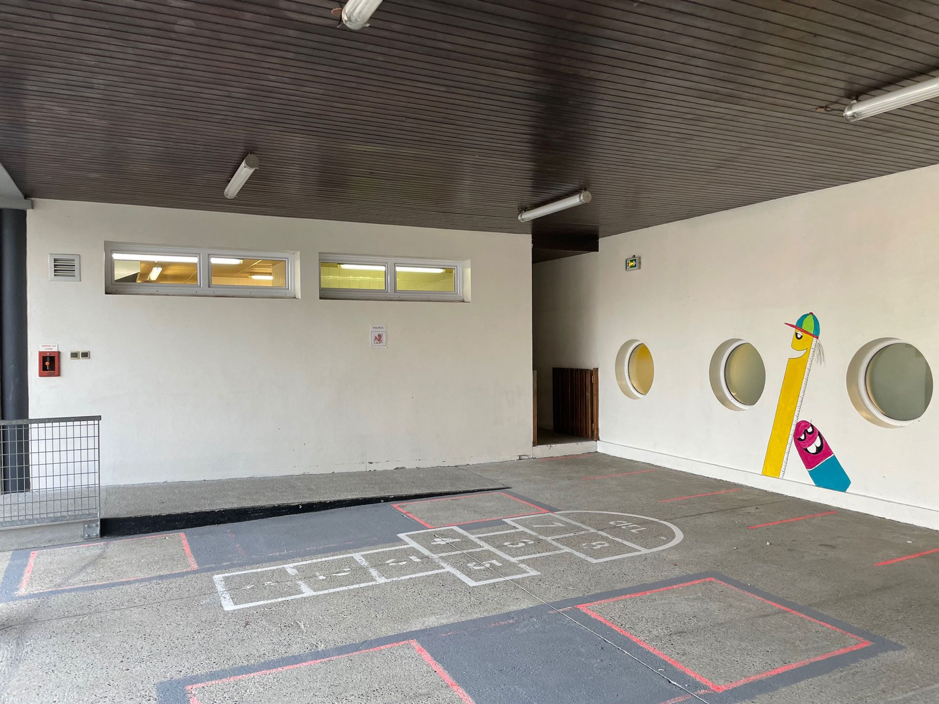 Une aire de jeux extérieure vide avec une marelle, des fenêtres circulaires et une fresque colorée sur un mur blanc.