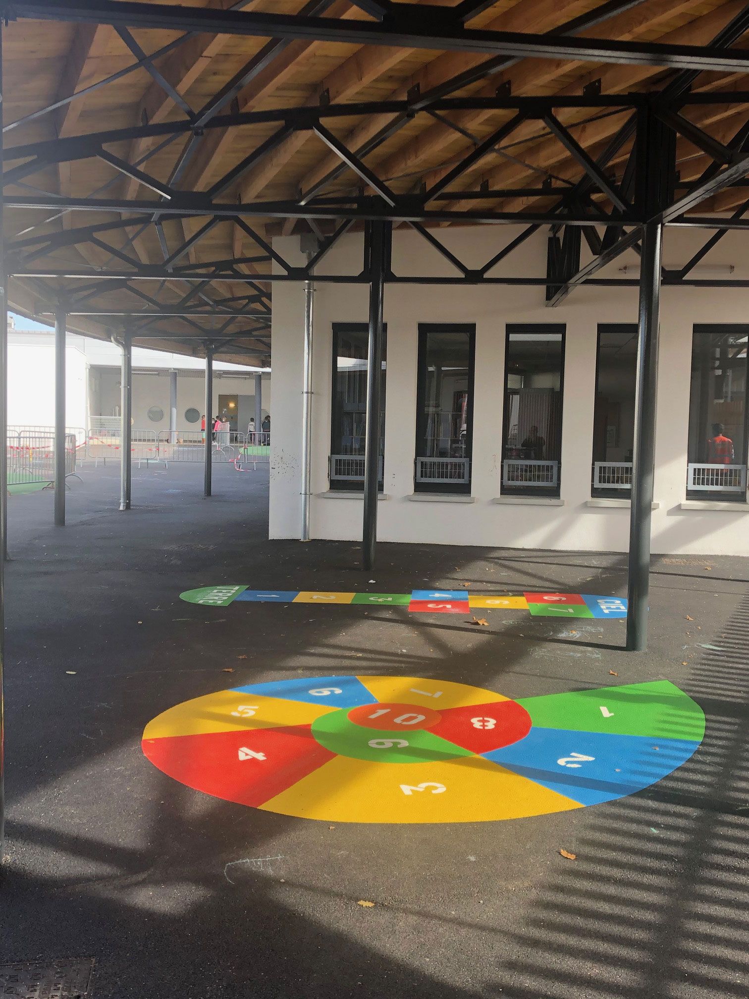 Aire de jeux avec une marelle colorée peinte au sol, sous une structure ombragée.