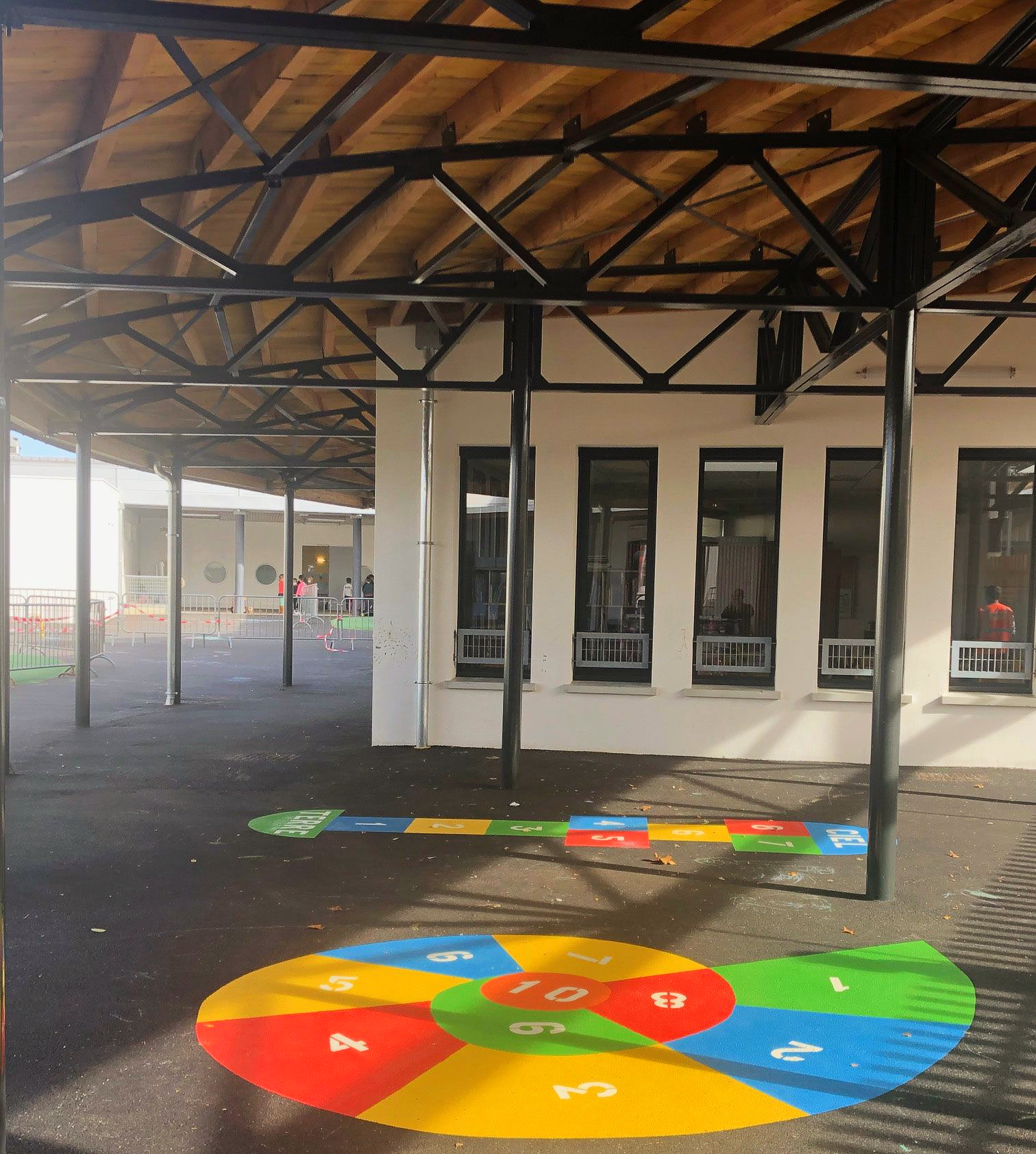 Aire de jeux avec une marelle colorée peinte au sol, sous une structure ombragée.