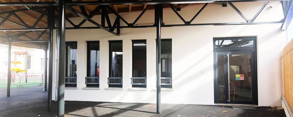 Vue extérieure d'un bâtiment blanc avec des fenêtres et une porte à encadrement noir. Passage couvert avec poutres métalliques au-dessus.