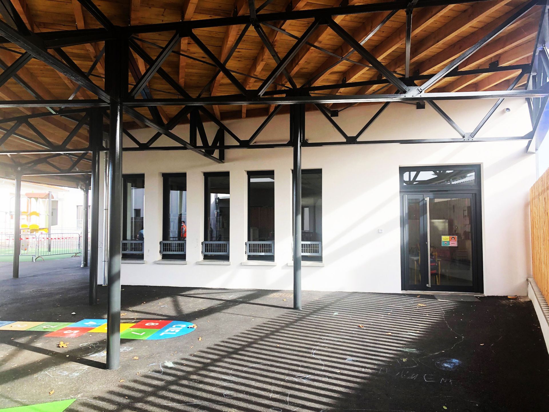 Espace extérieur couvert avec supports noirs, murs blancs, fenêtres, porte et plafond en bois apparent.