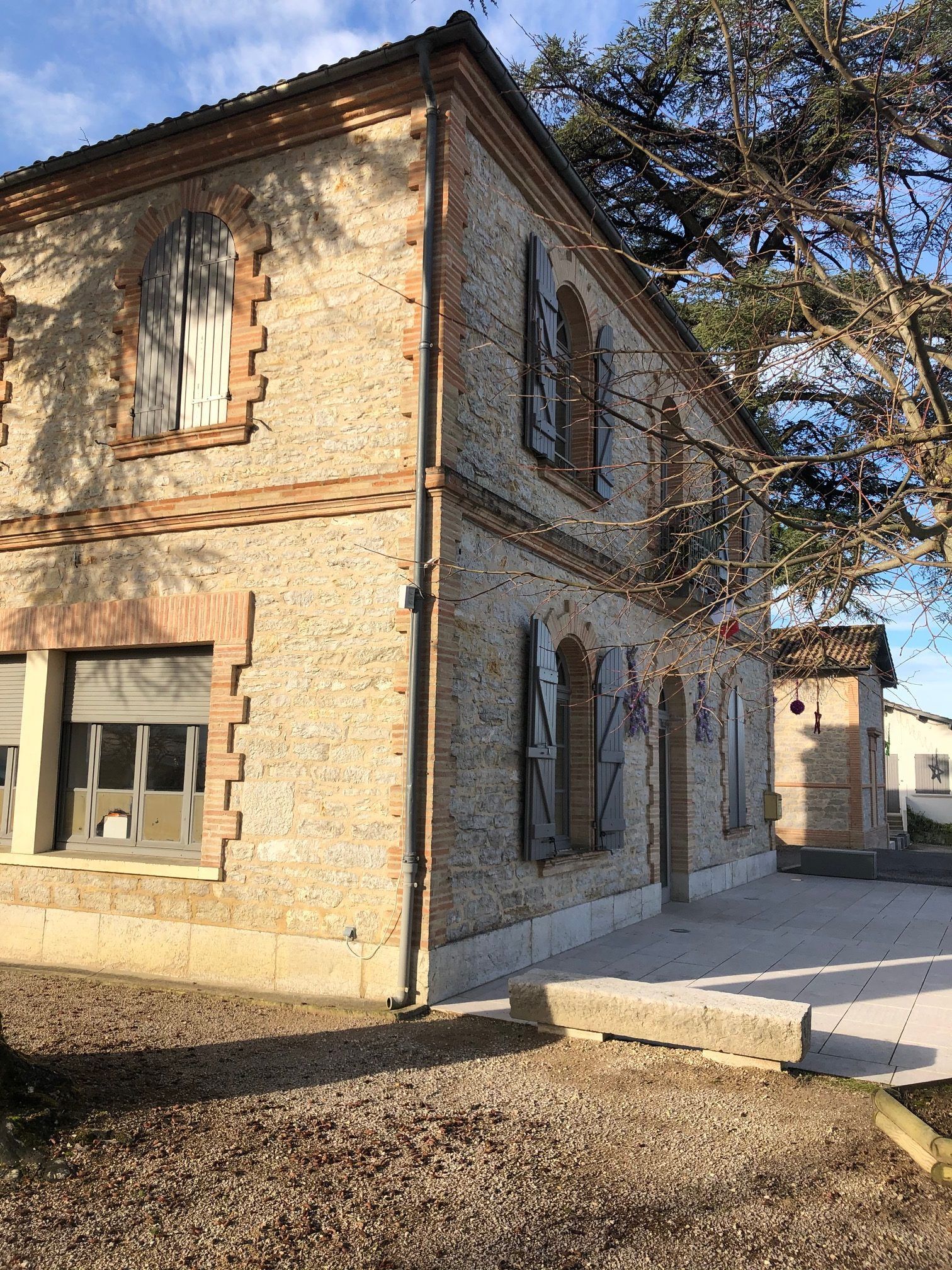 Bâtiment en briques de deux étages avec des éléments en pierre, volets fermés et un arbre par une journée ensoleillée.