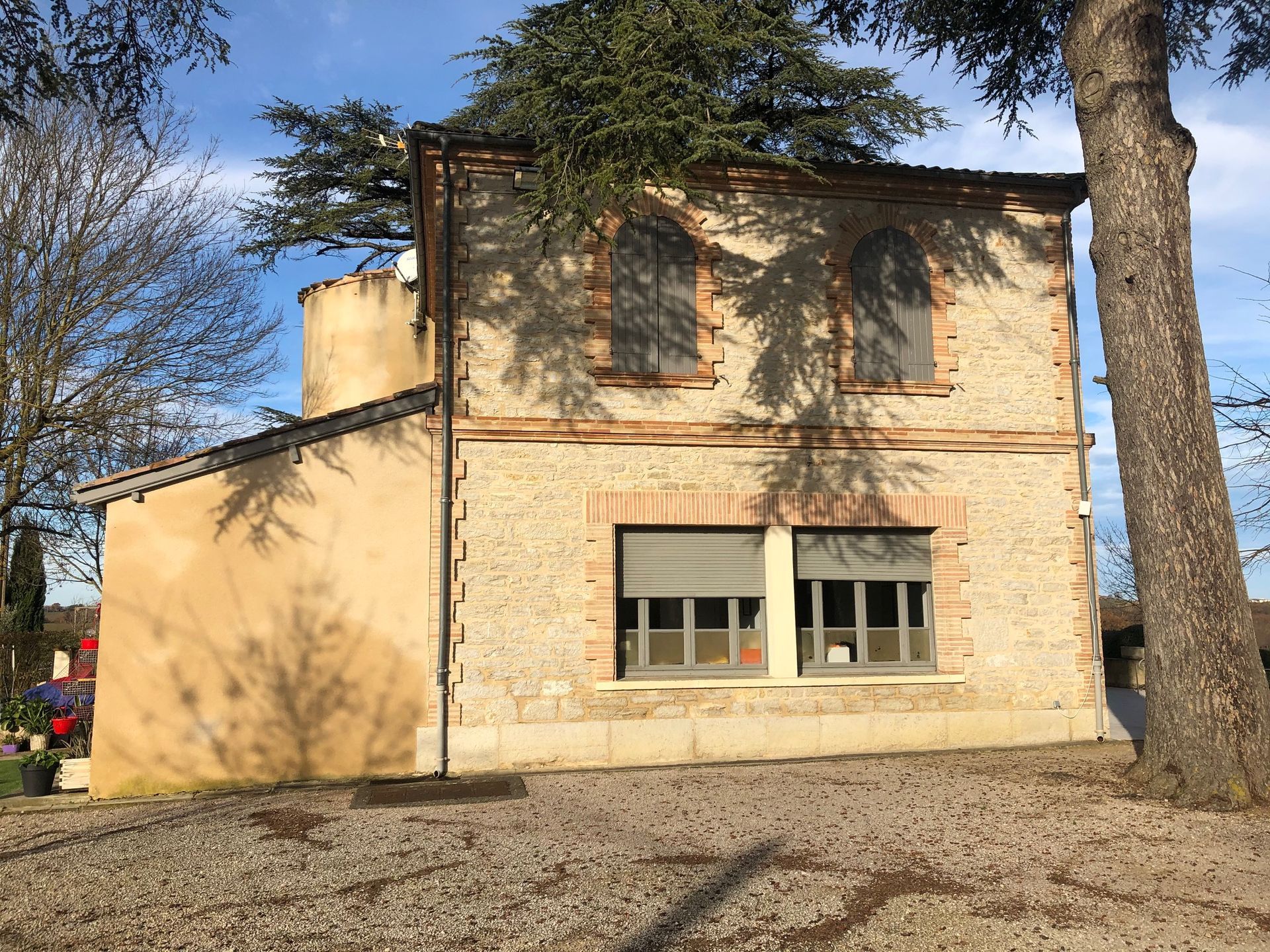 Maison en pierre et en brique, volets fermés, arbre au premier plan.