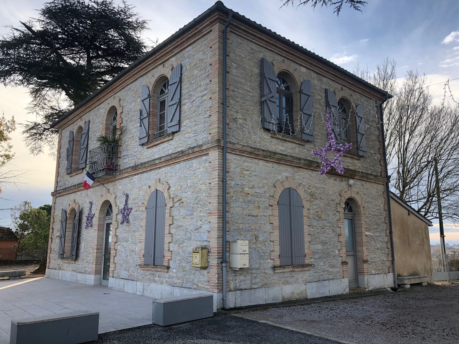 Bâtiment en pierre avec volets gris et drapeau français ; entouré d'arbres.