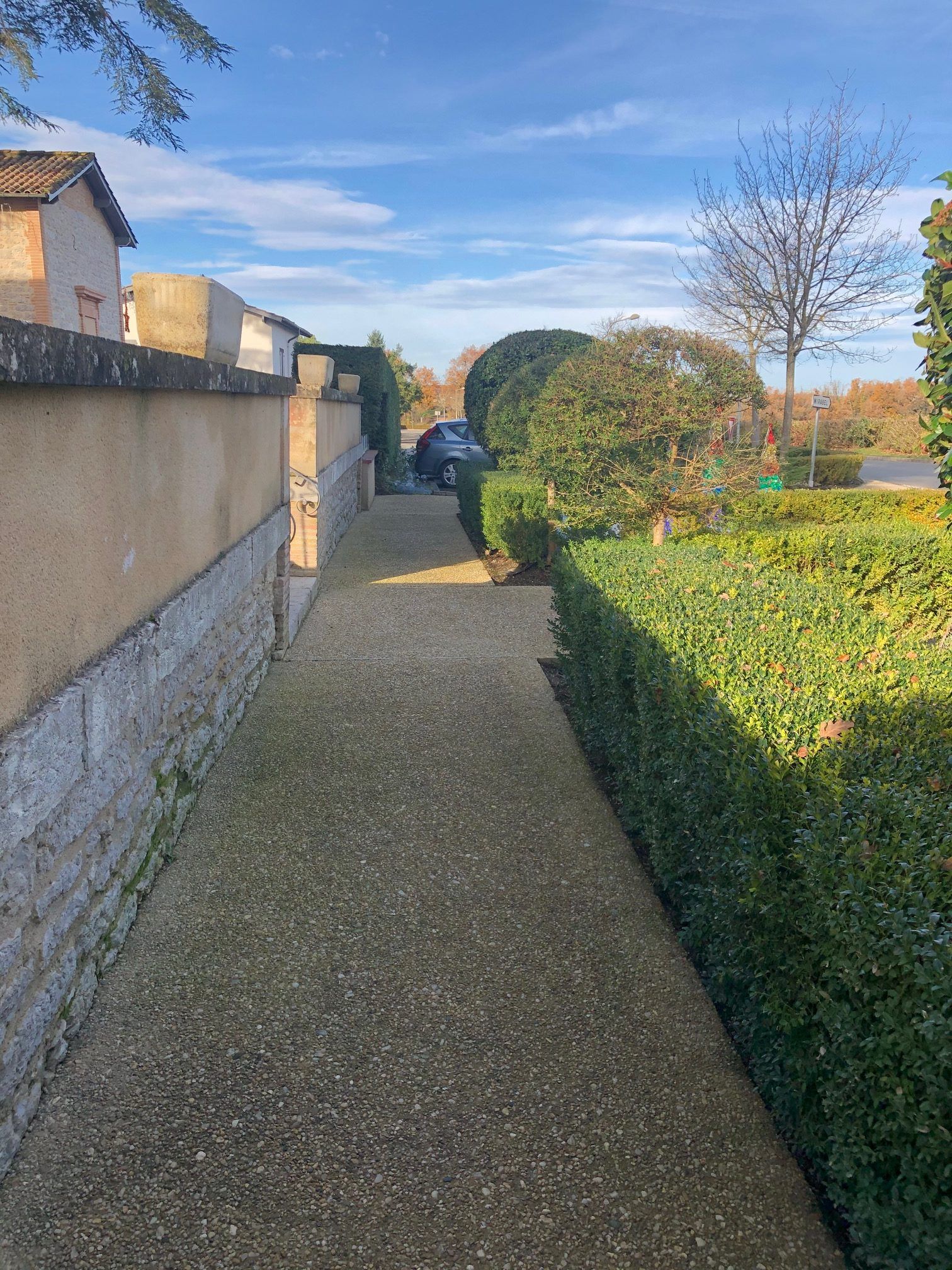 Un étroit chemin bordé de haies et de murs mène à une voiture et à des arbres sous un ciel bleu.