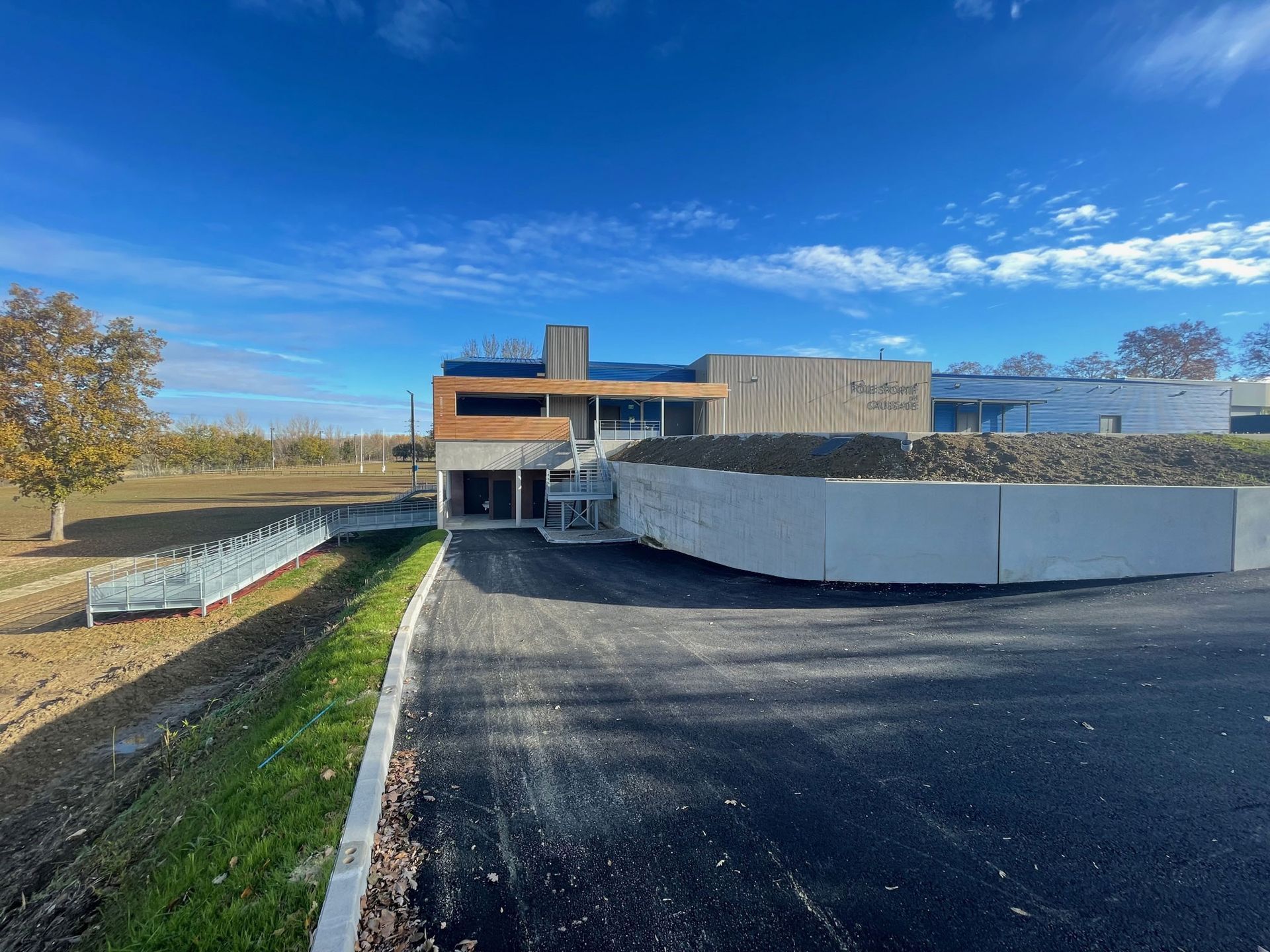 Bâtiment moderne sous un ciel bleu, allée asphaltée, façade en béton et en bois, quelques aménagements paysagers et un petit pont.