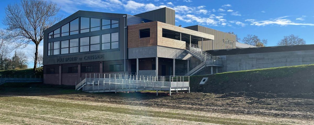 Un bâtiment moderne avec de grandes fenêtres et une façade en bois, jouxtant un espace vert sous un ciel bleu.