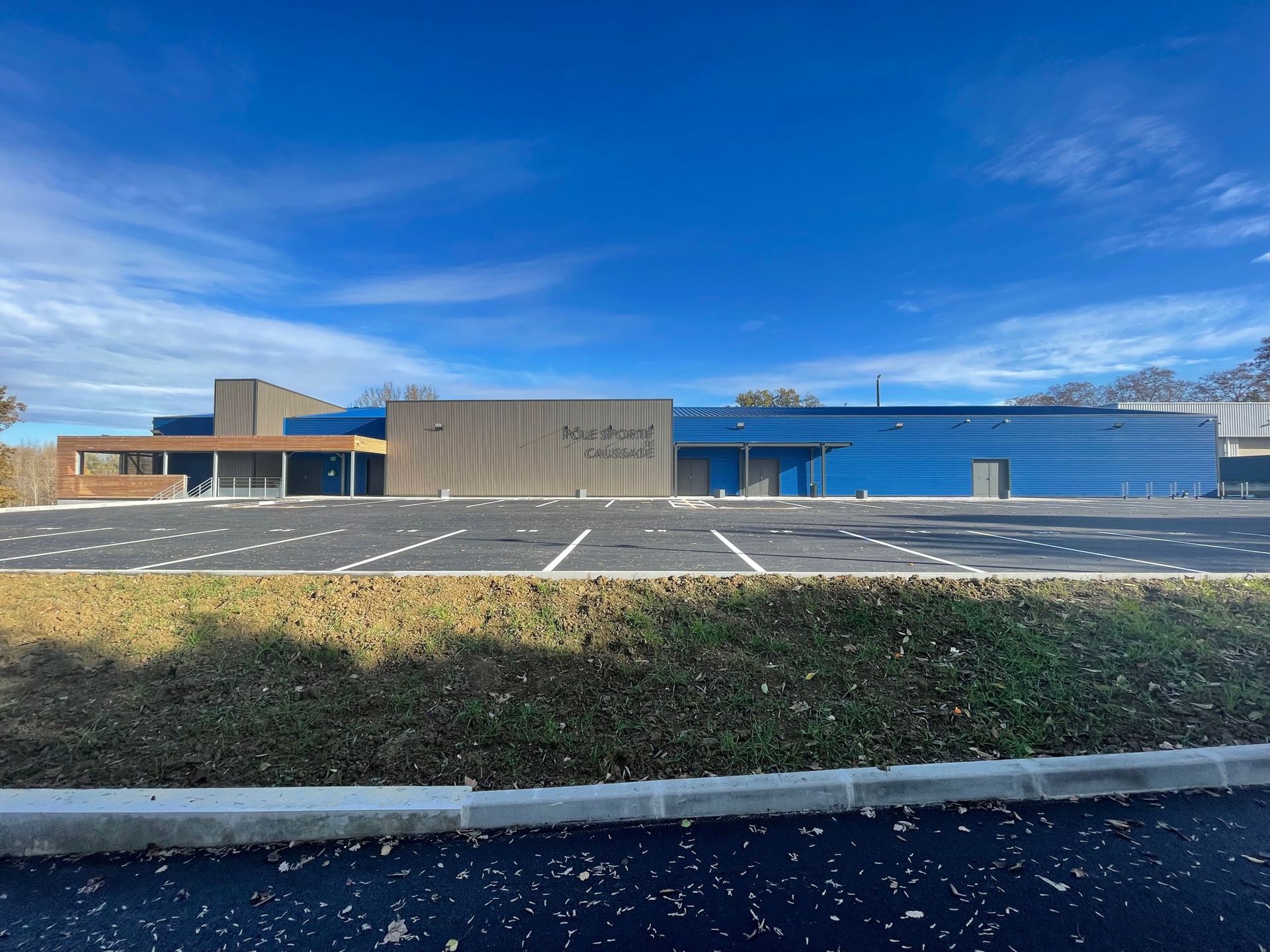 Vue extérieure d'un bâtiment moderne à façade bleue, avec des éléments en bois et un parking, sur fond de ciel bleu éclatant.
