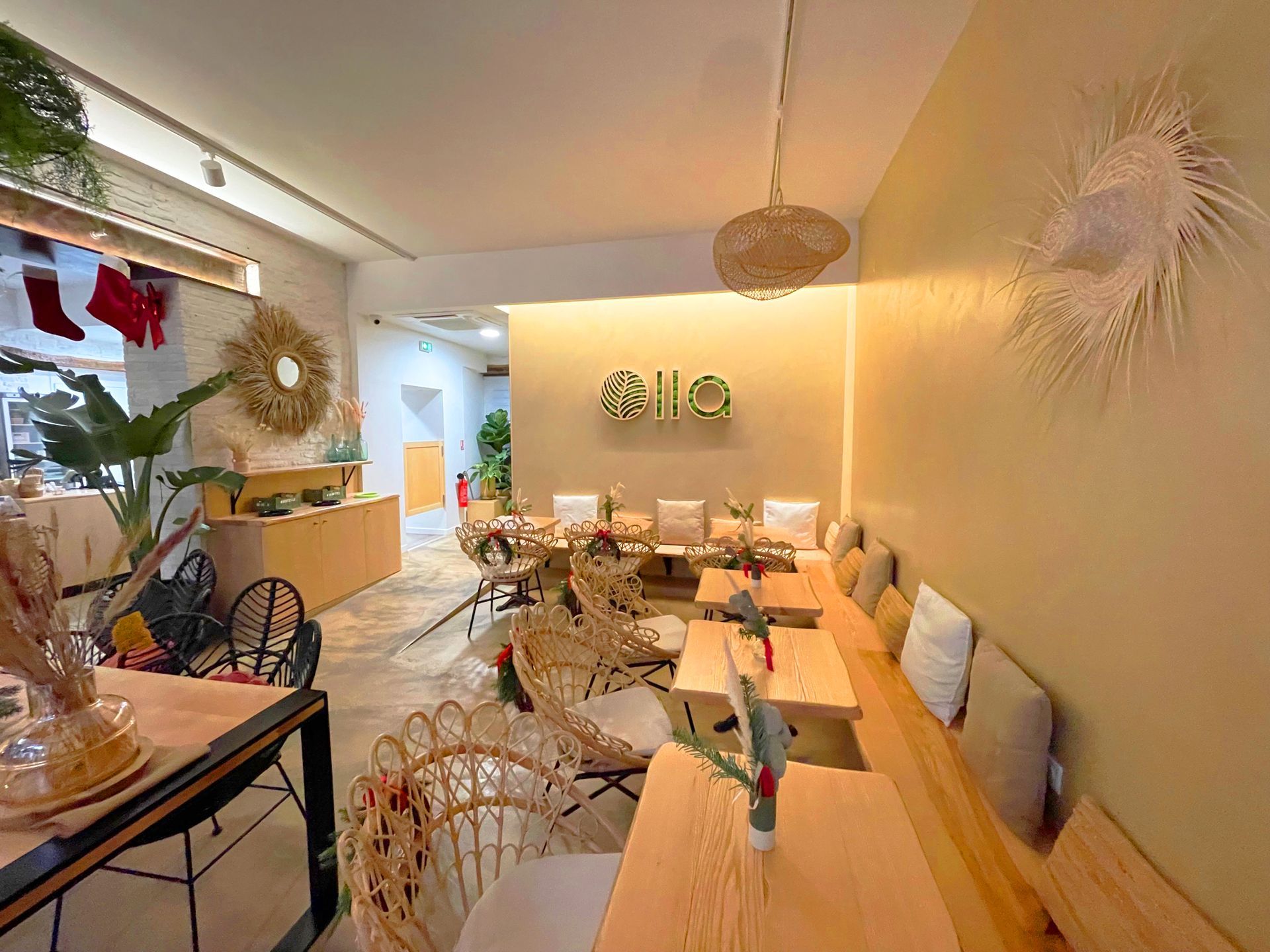 Intérieur d'un café avec chaises en rotin, tables en bois et banquette avec coussins. Lumière naturelle, plantes et décoration.