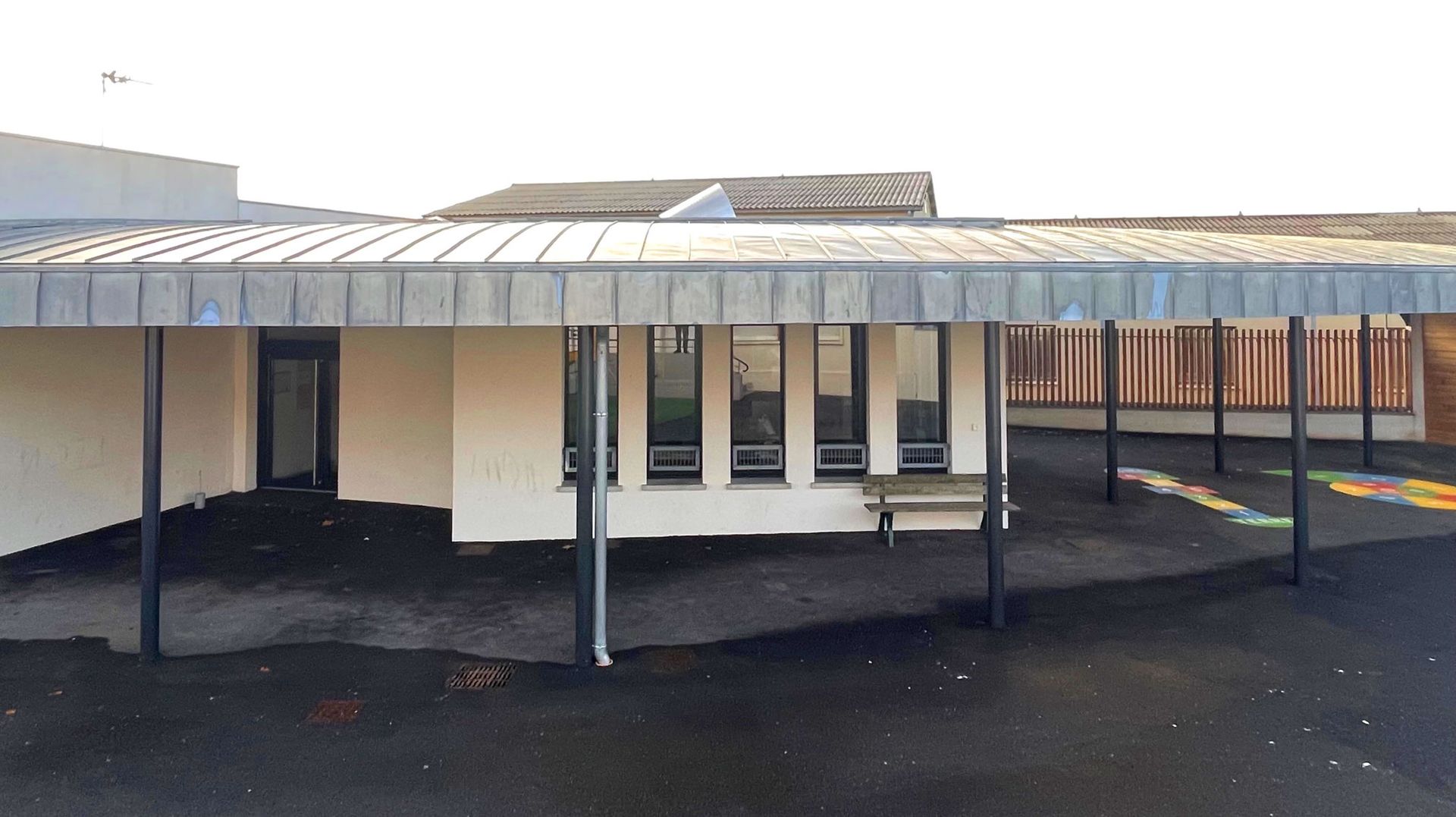Extérieur de l'école avec allée couverte ; murs blancs, toit en métal, sol noir.