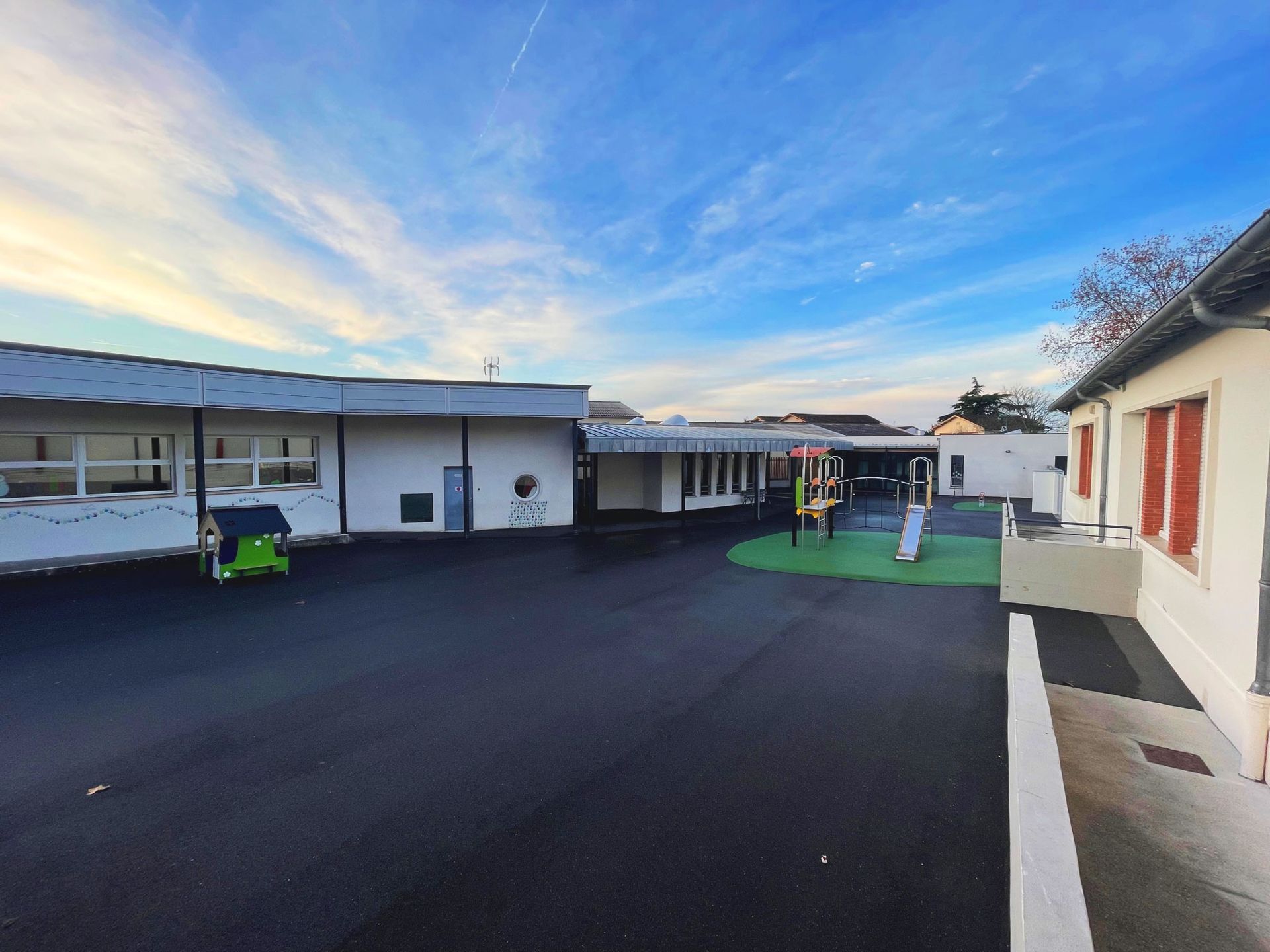 Cour d'école avec bâtiments, jeux pour enfants et ciel couvert.