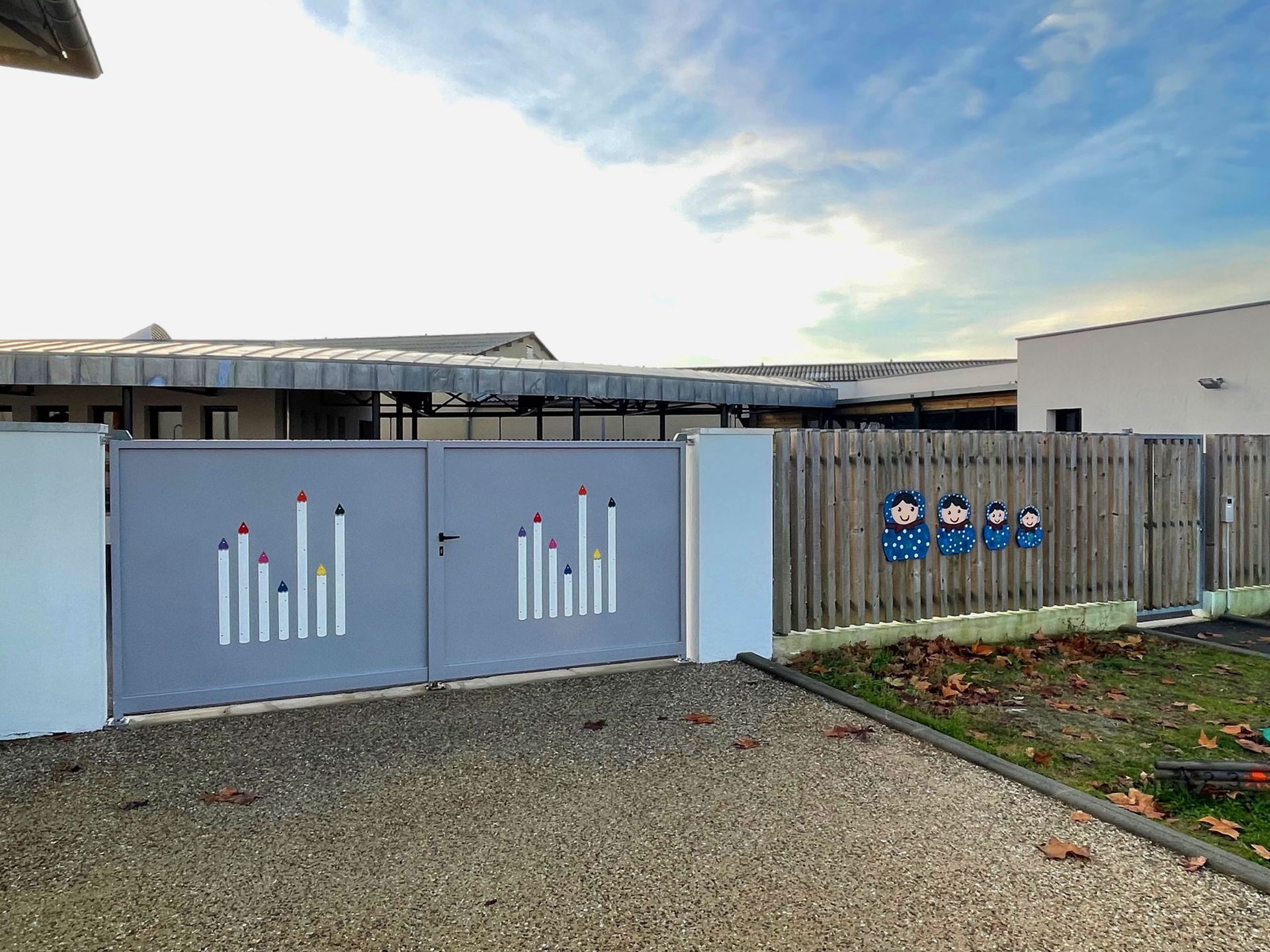 Un portail gris orné de motifs stylisés évoquant des bougies donne accès à un bâtiment. La clôture en bois adjacente est décorée sur le thème de l'enfance.