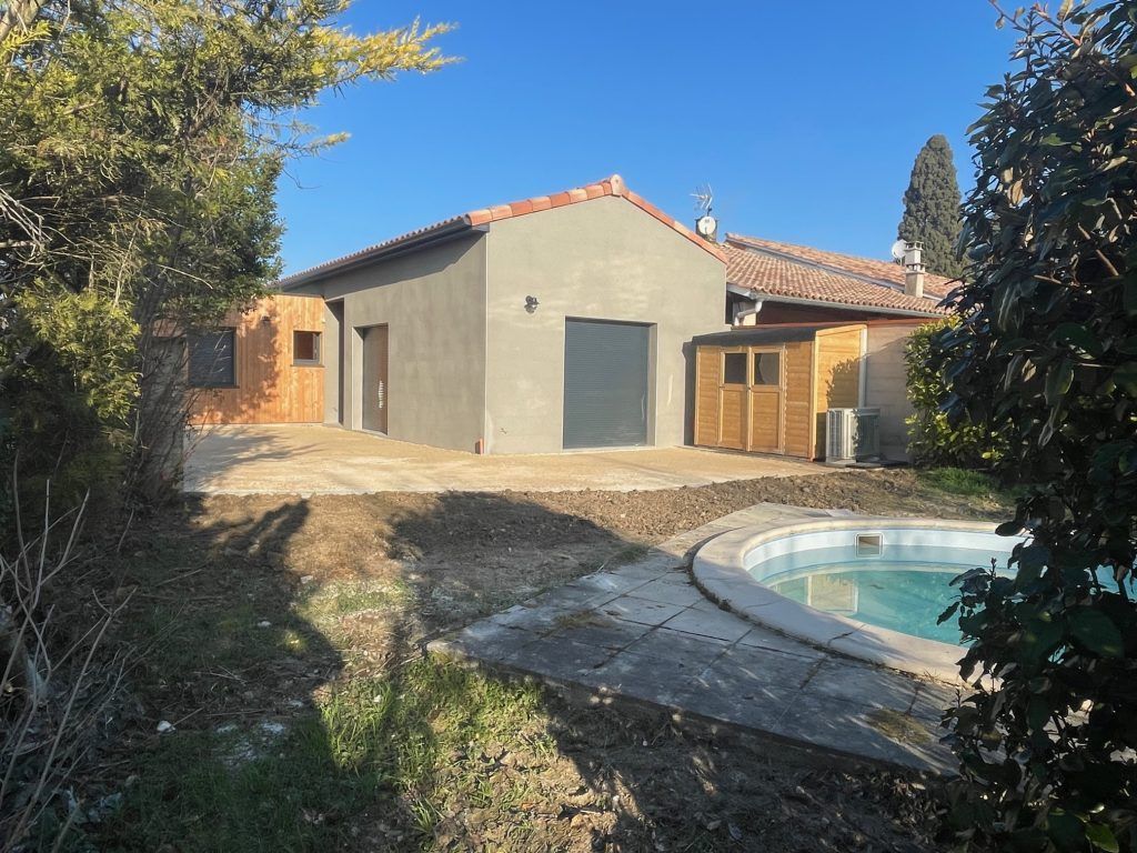 Maison avec piscine. La façade est grise, le toit en tuiles rouges et il y a un abri de jardin en bois. Journée ensoleillée.