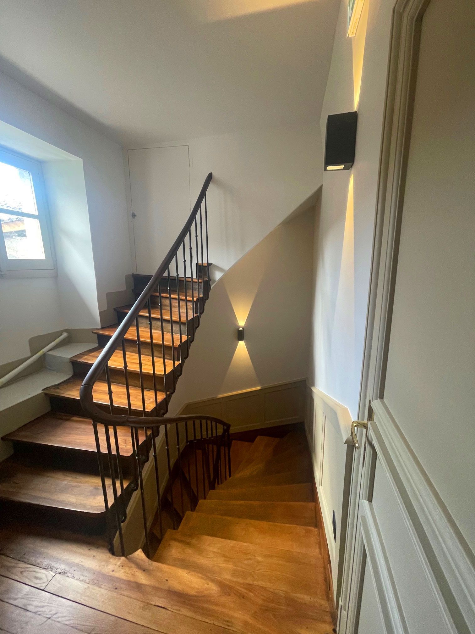 Escalier en bois dans un bâtiment avec une base incurvée, une rampe noire et des appliques murales.