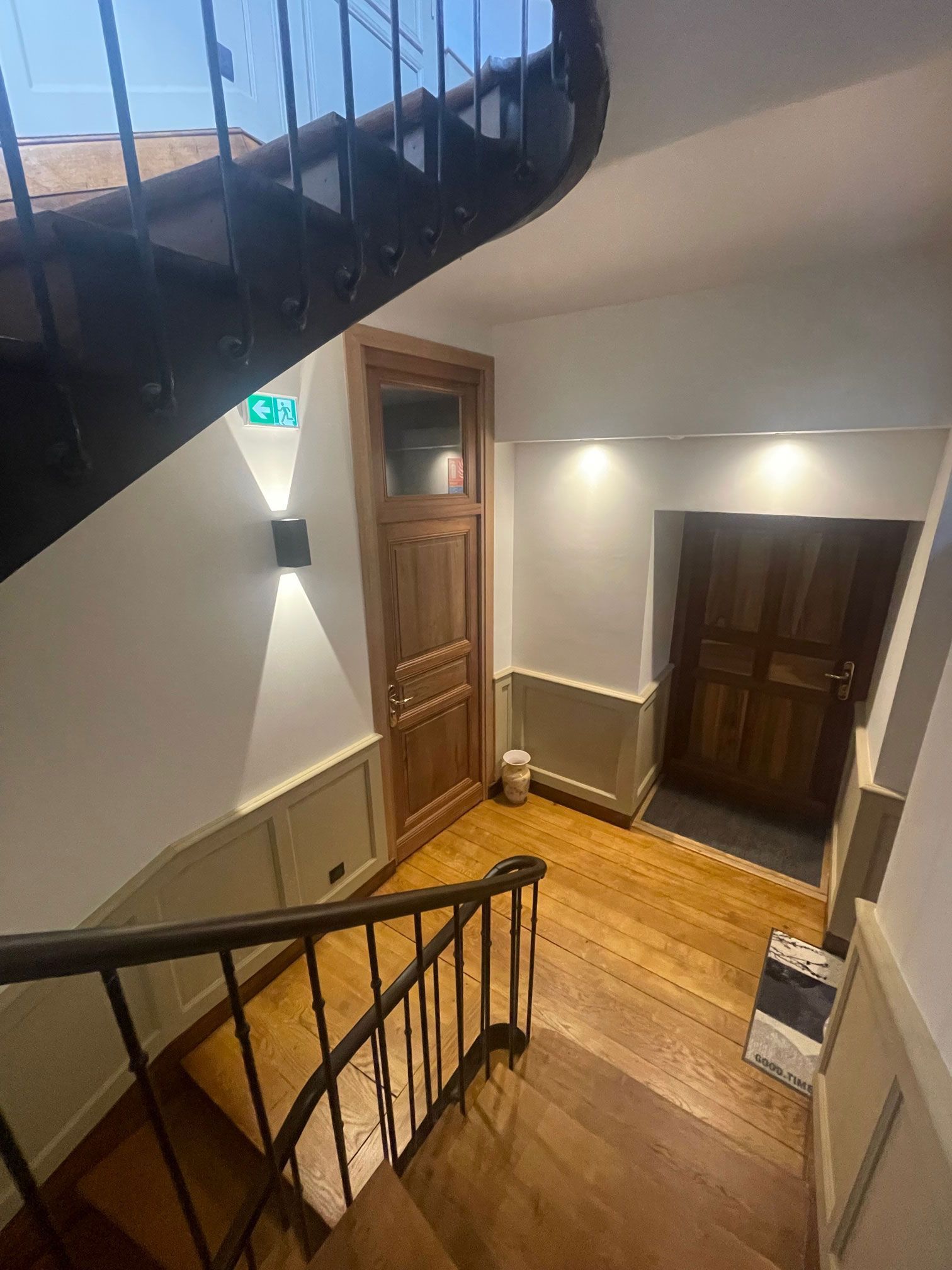 Escalier en bois avec rampe noire et deux portes en bois. Murs blancs et éclairage encastré.