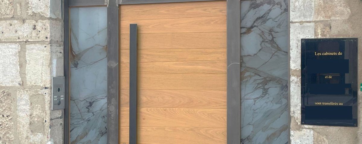 Porte en bois à cadre métallique, encastrée dans un mur en pierre. Une boîte aux lettres se trouve à côté de la porte.