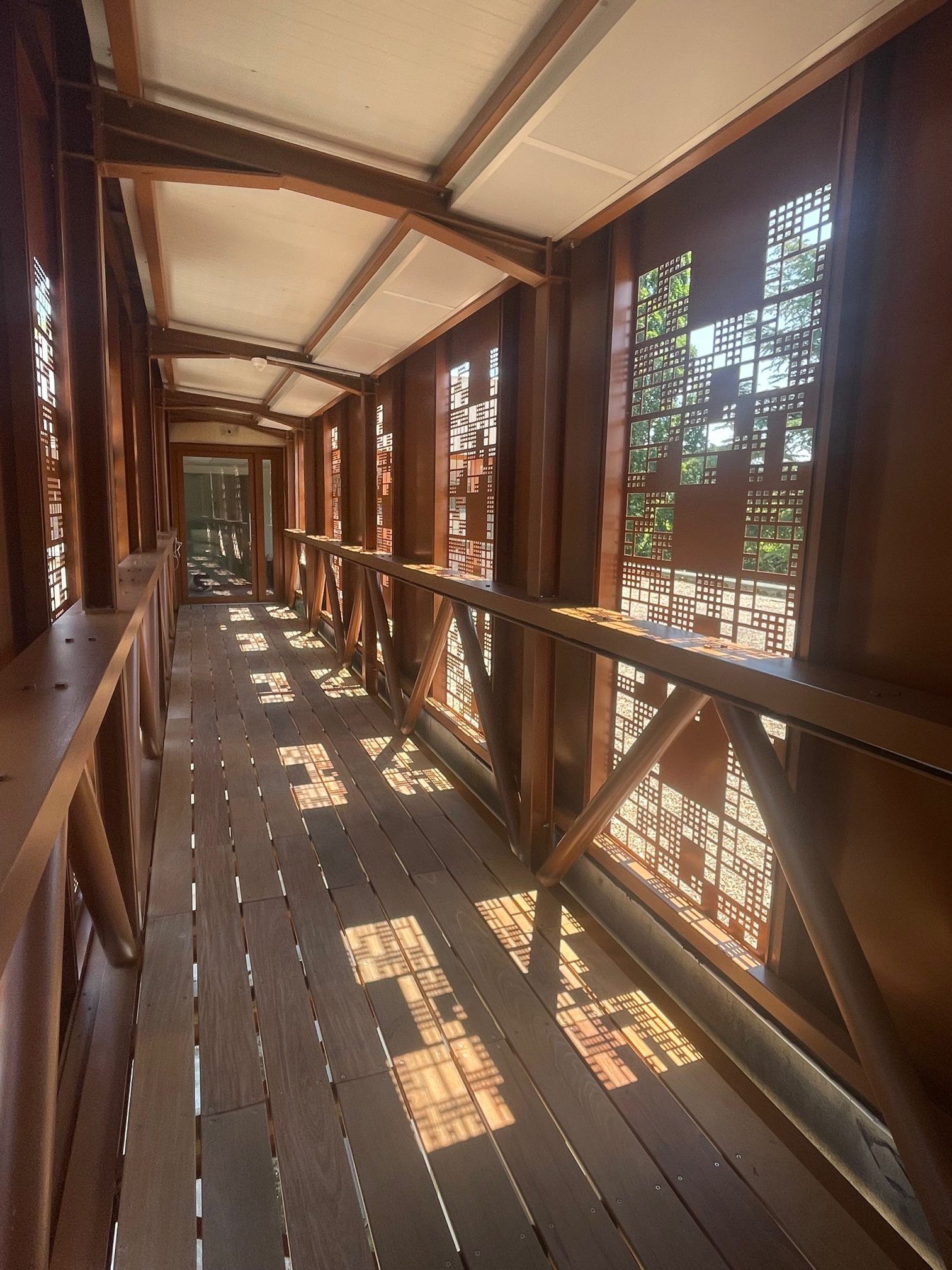 Passerelle en bois ornée de découpes décoratives, menant à une zone verdoyante floue ; la lumière du soleil filtre à travers les ouvertures.