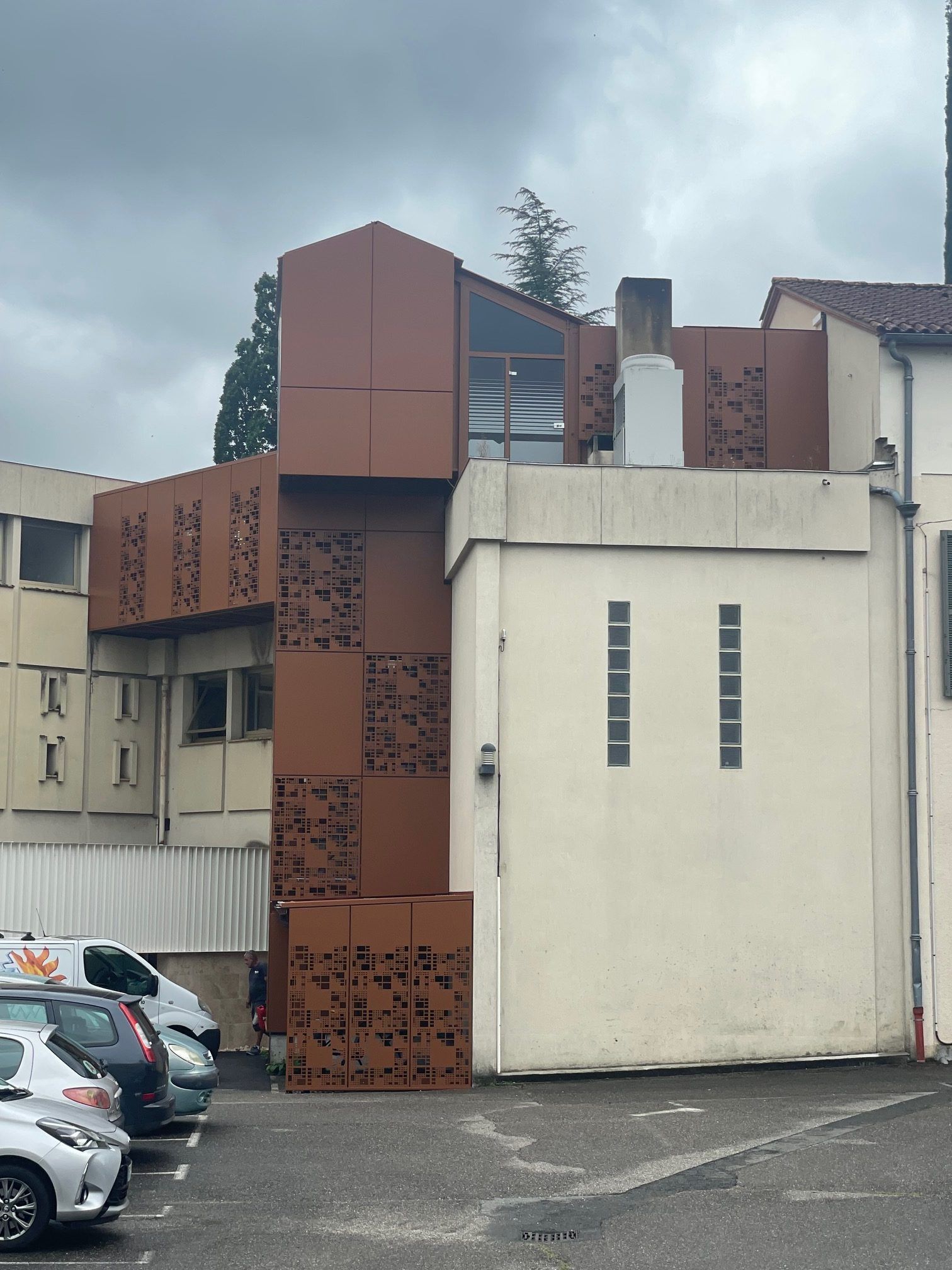 Façade métallique couleur rouille d'un bâtiment comportant des parties blanches et beiges ; voitures garées au premier plan sous un ciel couvert.