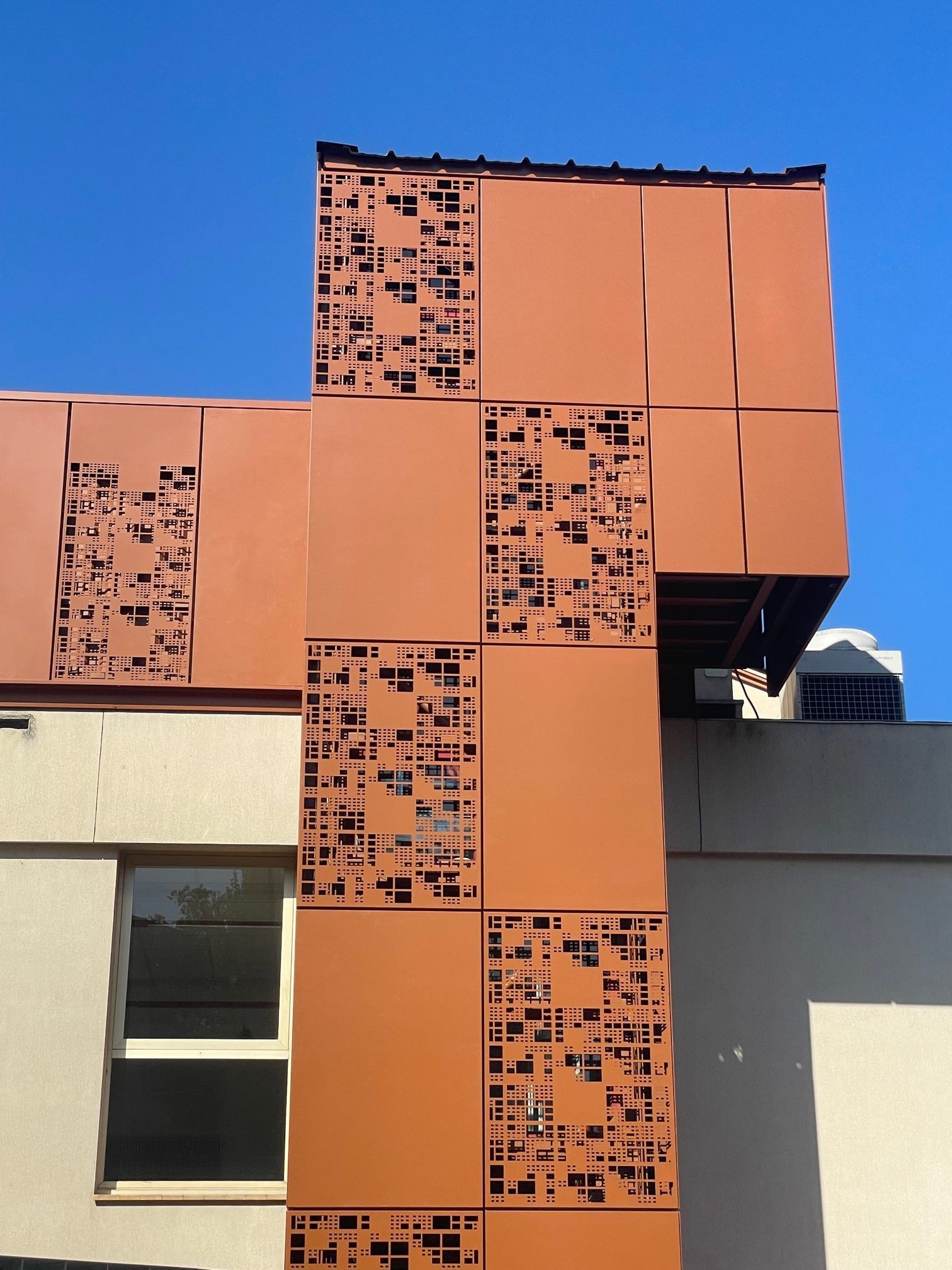 Façade métallique orange ajourée sur un bâtiment se détachant sur un ciel bleu clair.