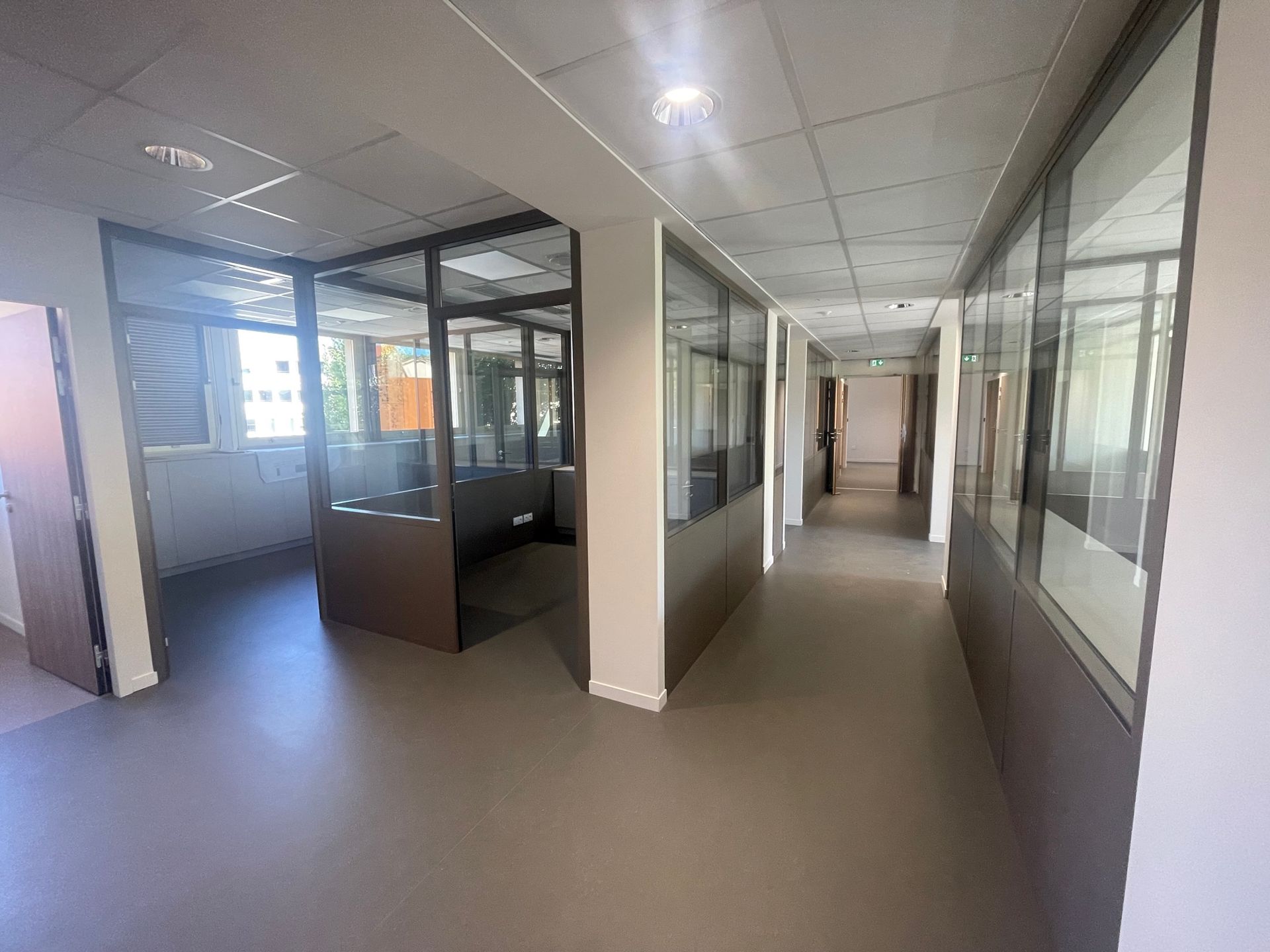 Couloir intérieur de bureaux avec pièces et portes vitrées. Sol gris, murs neutres, éclairage fluorescent.