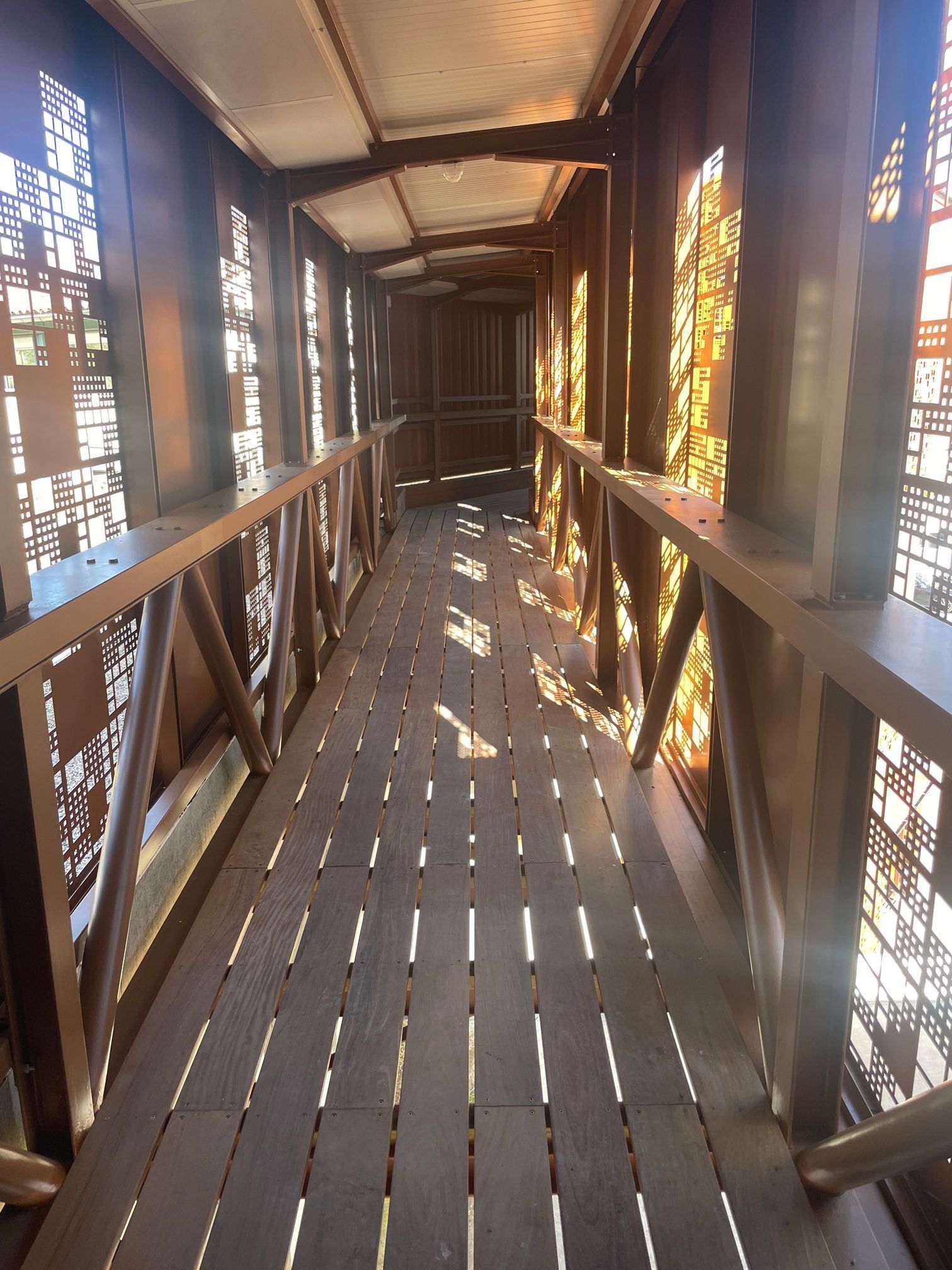 Passerelle métallique avec panneaux latéraux à motifs et poutres supérieures. La lumière du soleil projette des ombres sur le plancher en bois.