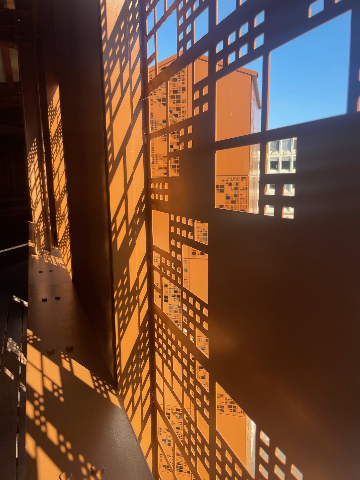 Façade métallique couleur rouille, ornée de découpes géométriques projetant des ombres. Ciel bleu visible à travers les ouvertures.