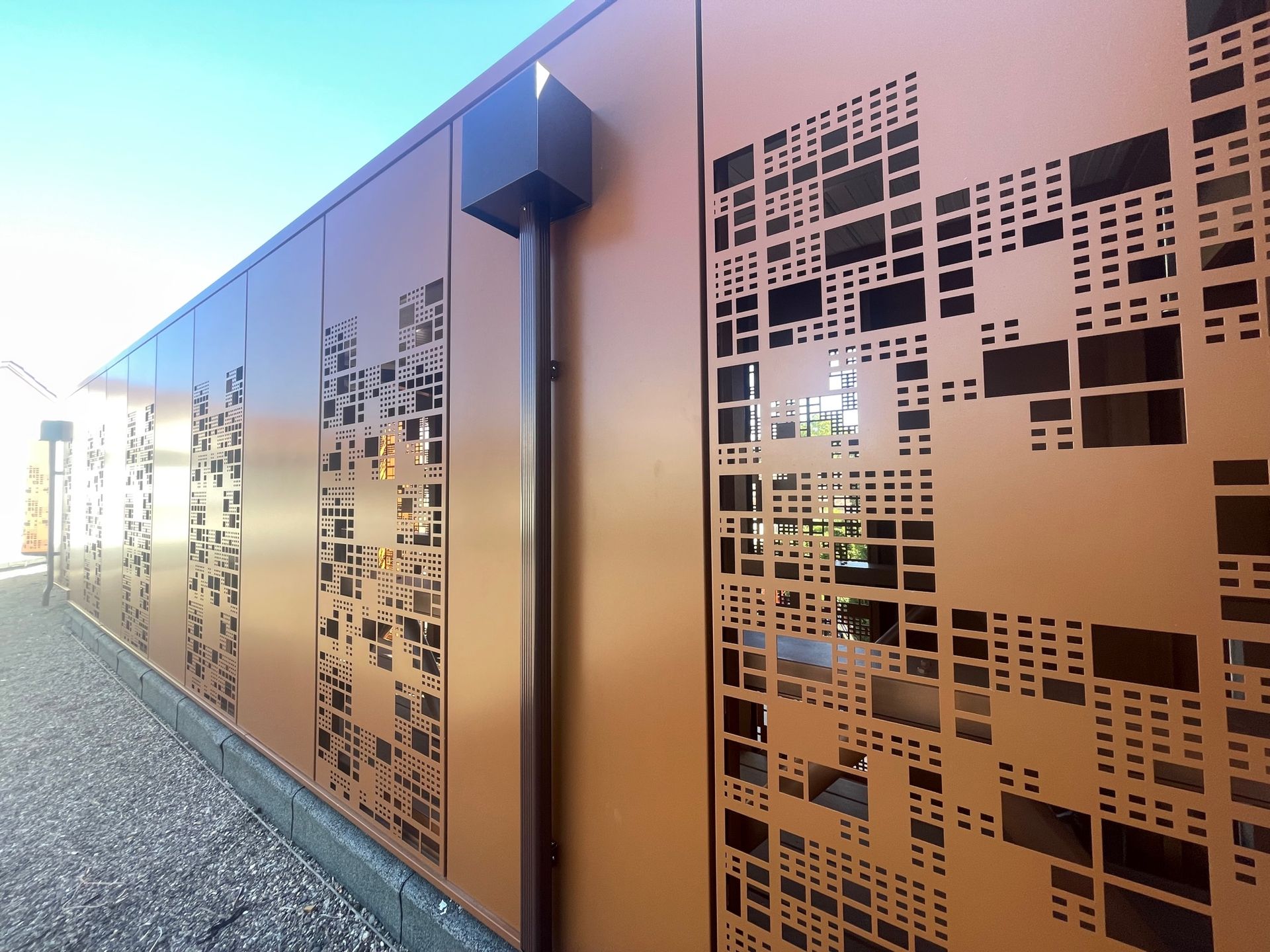 Mur en métal brun avec découpes géométriques et descente de gouttière, sur fond de ciel bleu.