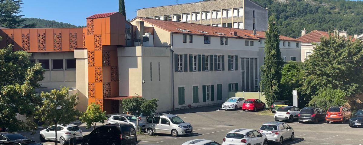 Bâtiments à façade métallique rouillée et parking avec des voitures. Montagne en arrière-plan. Journée ensoleillée.