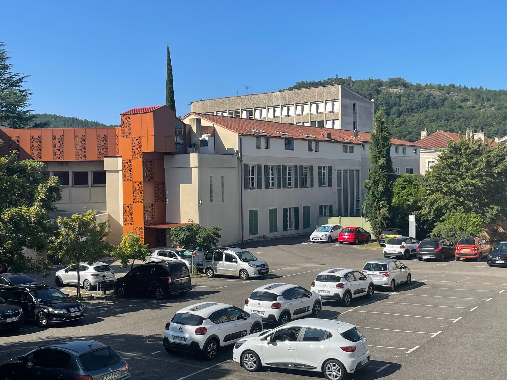Parking avec des voitures devant un bâtiment clair sous un ciel bleu, montagnes en arrière-plan.