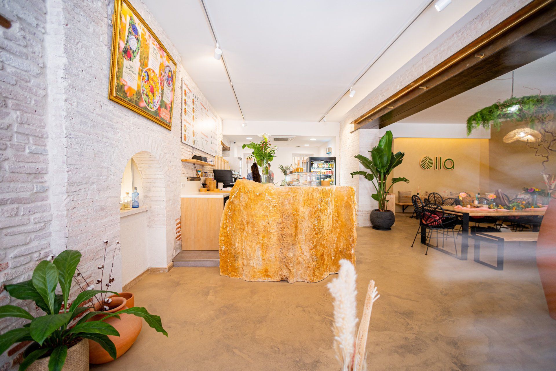 Intérieur lumineux de café avec des murs blancs texturés, un comptoir d'accueil doré et des plantes en pot.
