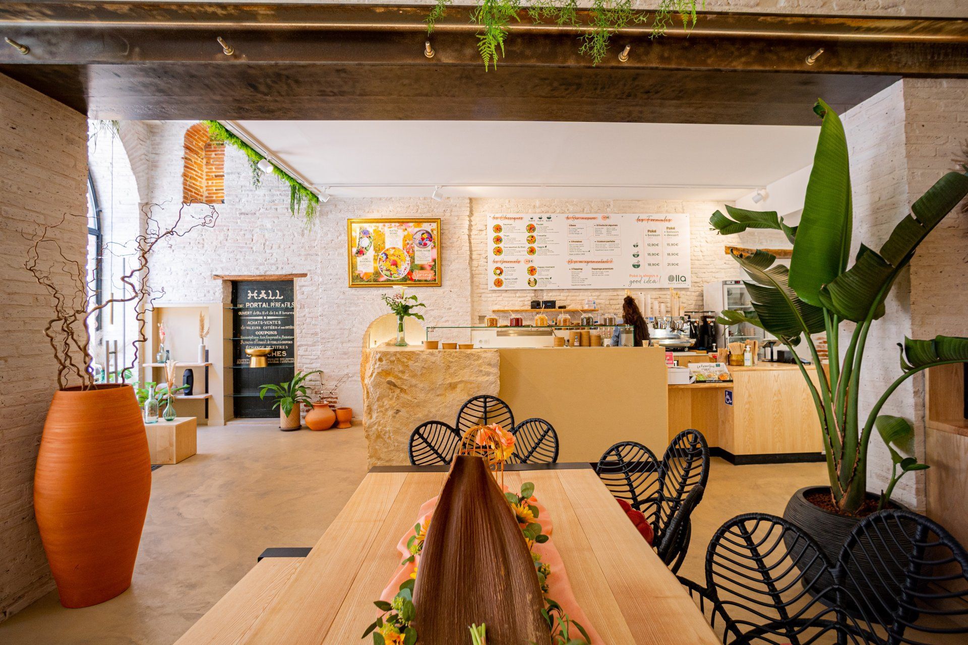 Intérieur d'un café avec une longue table en bois, de grandes plantes en pot et un mur de briques blanches.