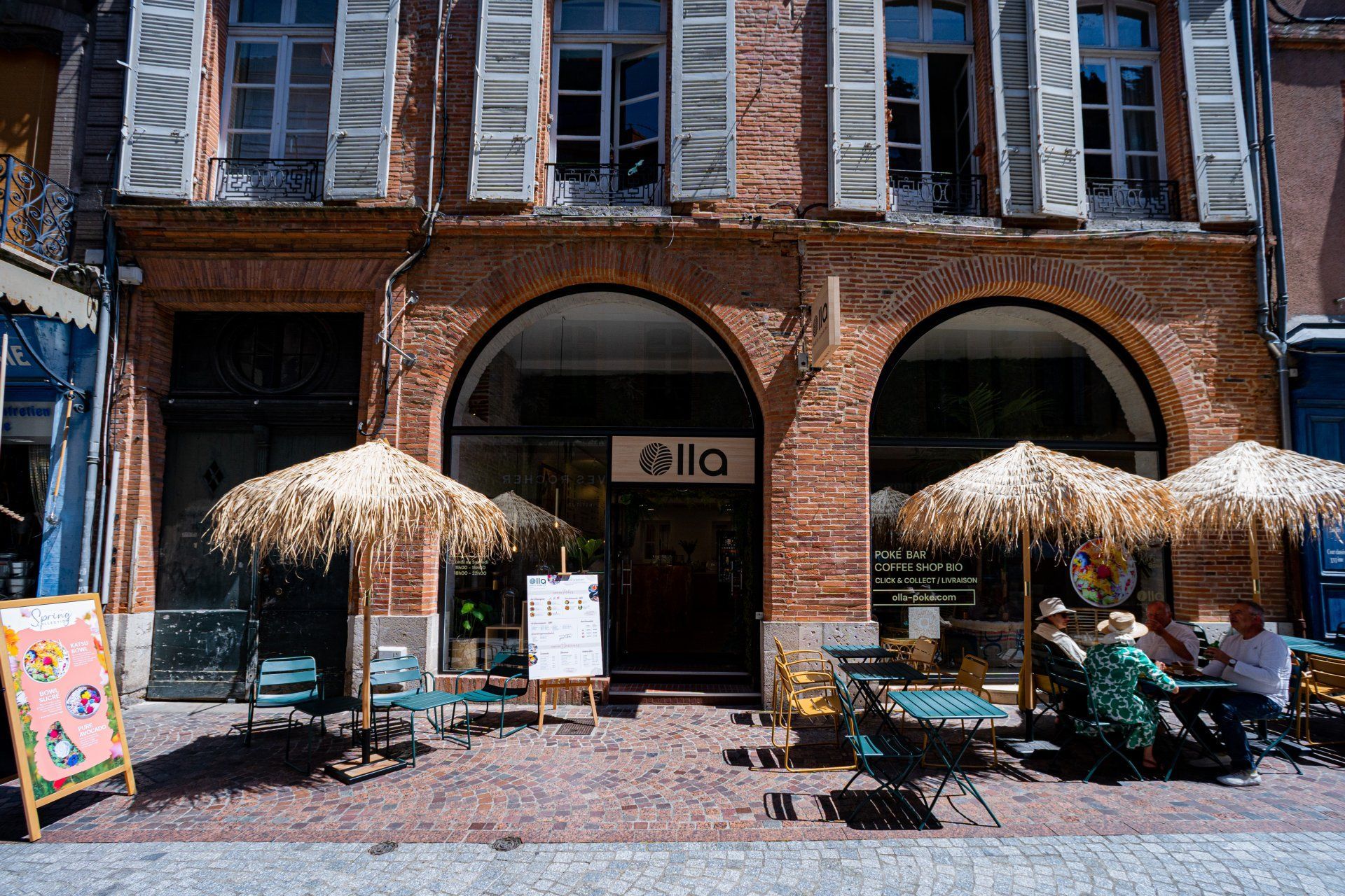 Extérieur d'un café avec parasols en paille, tables et chaises. Le bâtiment possède des fenêtres cintrées et une façade en briques.